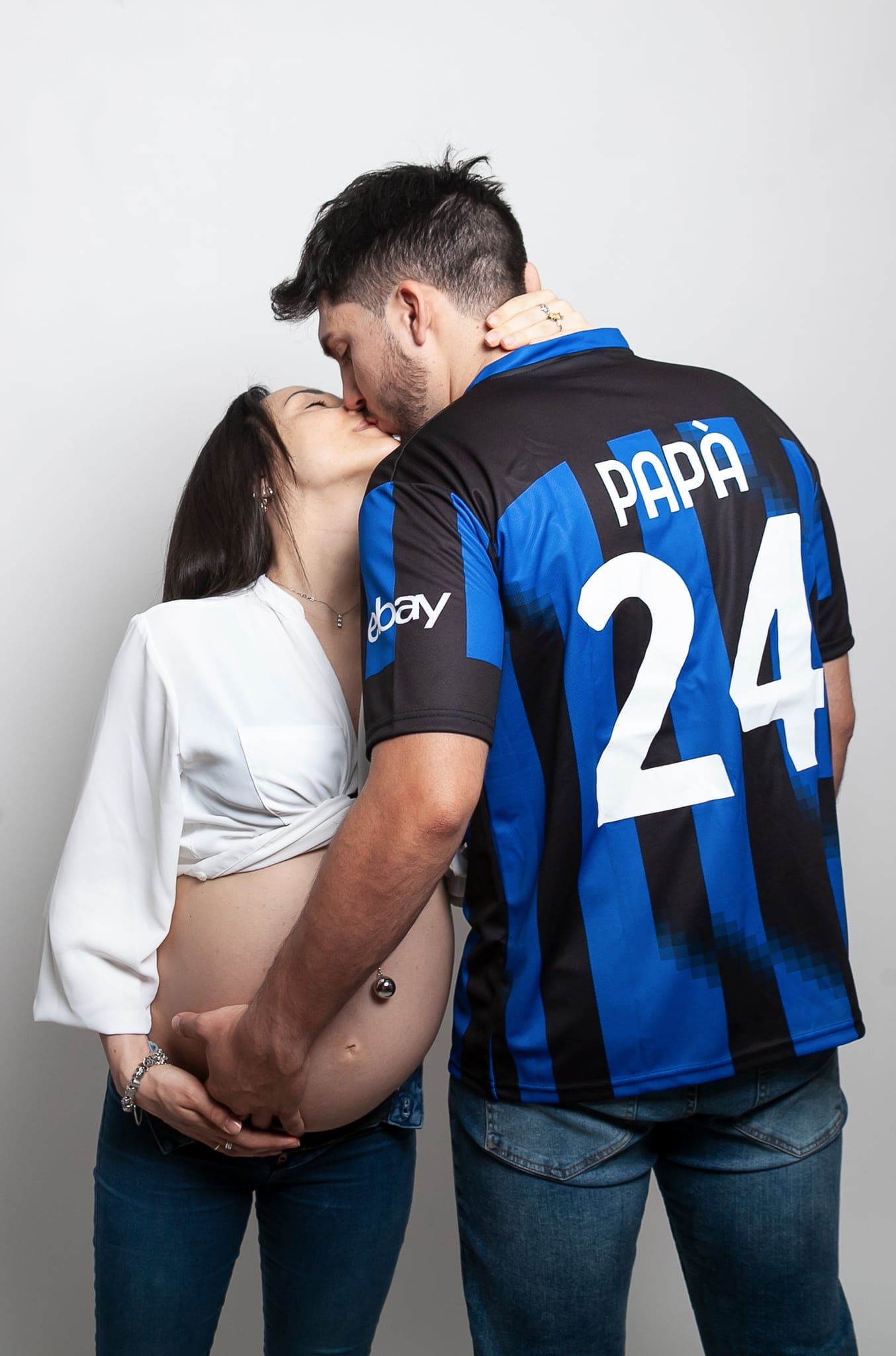 Futuri genitori che si baciano con le mani sul pancione, lui di spalle con la maglia blu e nera con scritto PAPÀ 24, su fondo chiaro.
