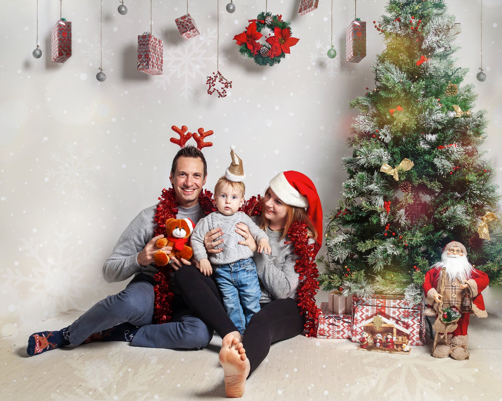 Mamma, papà e bambino piccolo seduti a terra con tinsel rosso e cerchietti natalizi, accanto all'albero e a un Babbo Natale decorativo