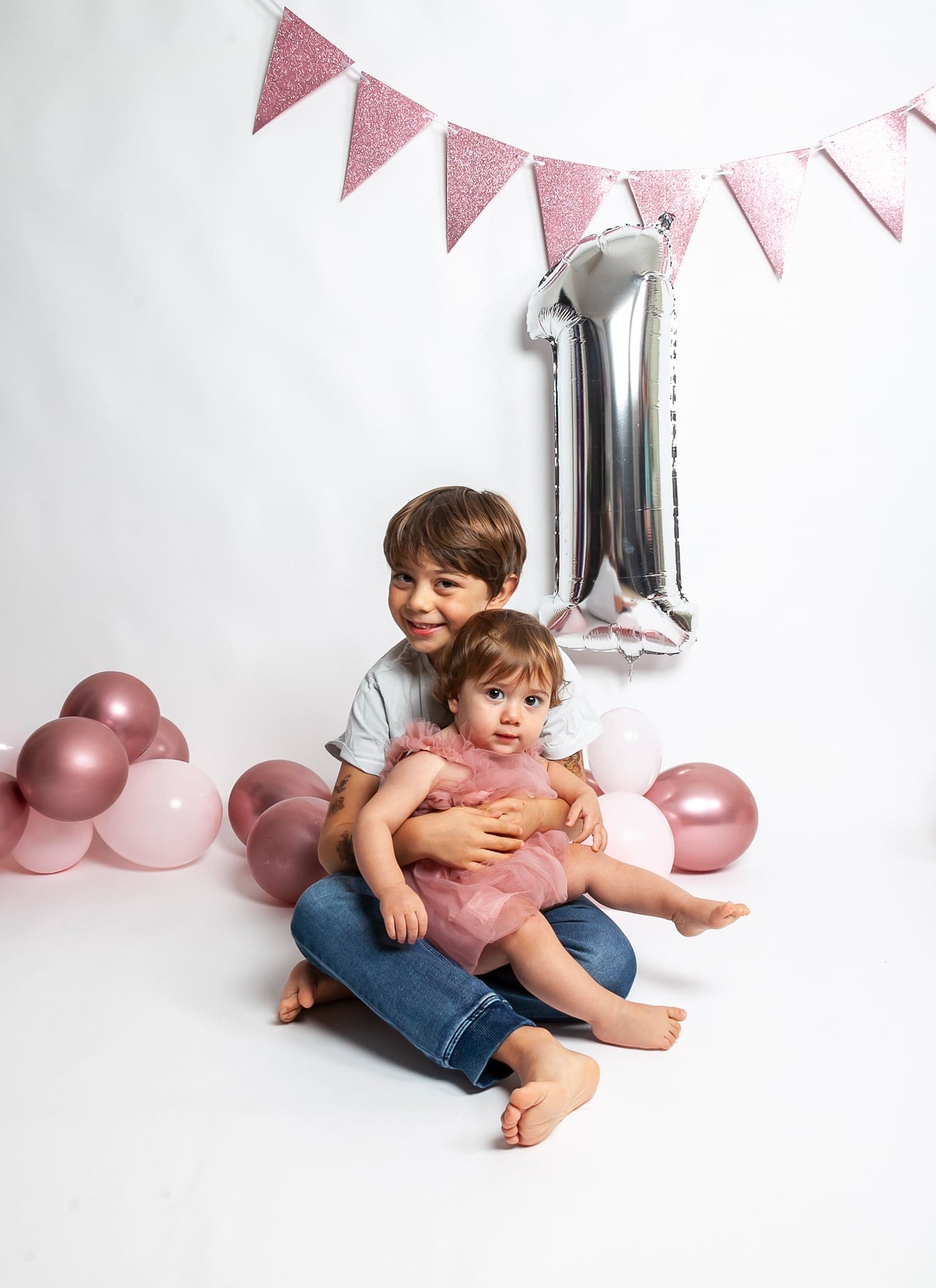 Un bambino sorride tenendo sulle gambe la sorellina in abito di tulle rosa, davanti a palloncino argentato e festone rosa