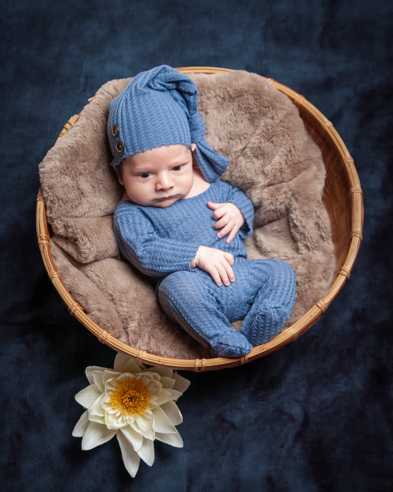 Neonato con cuffia e tutina di lana blu disteso in un cesto con pelliccia beige, su sfondo blu scuro con un fiore bianco.