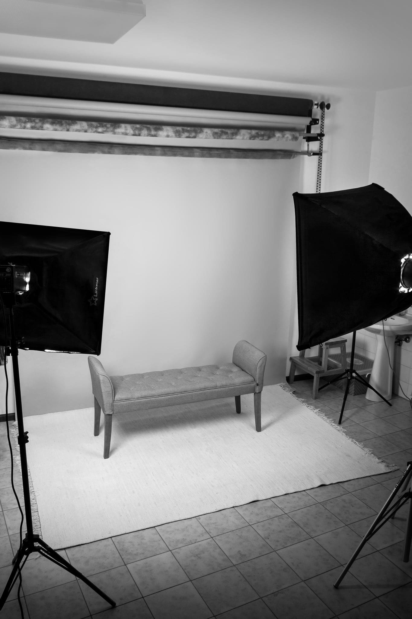 Studio fotografico con panca imbottita su tappeto chiaro, due softbox e fondale appeso, in bianco e nero