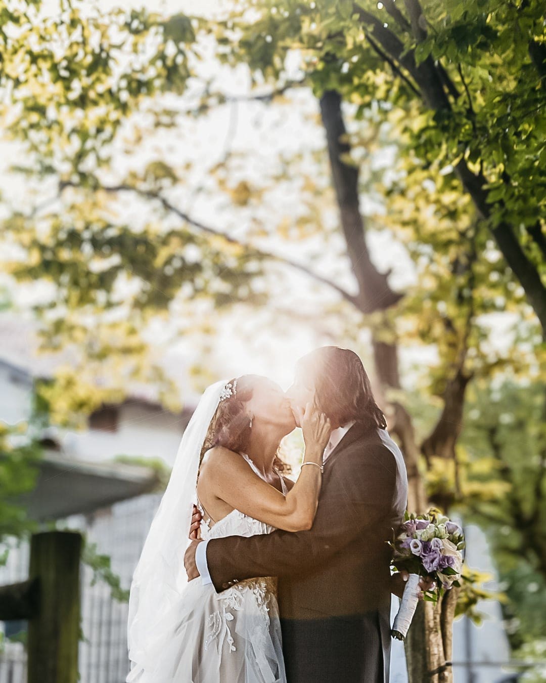 Sposi che si baciano in controluce tra gli alberi, con il bouquet viola, durante il matrimonio a Pagnacco, vicino a Udine
