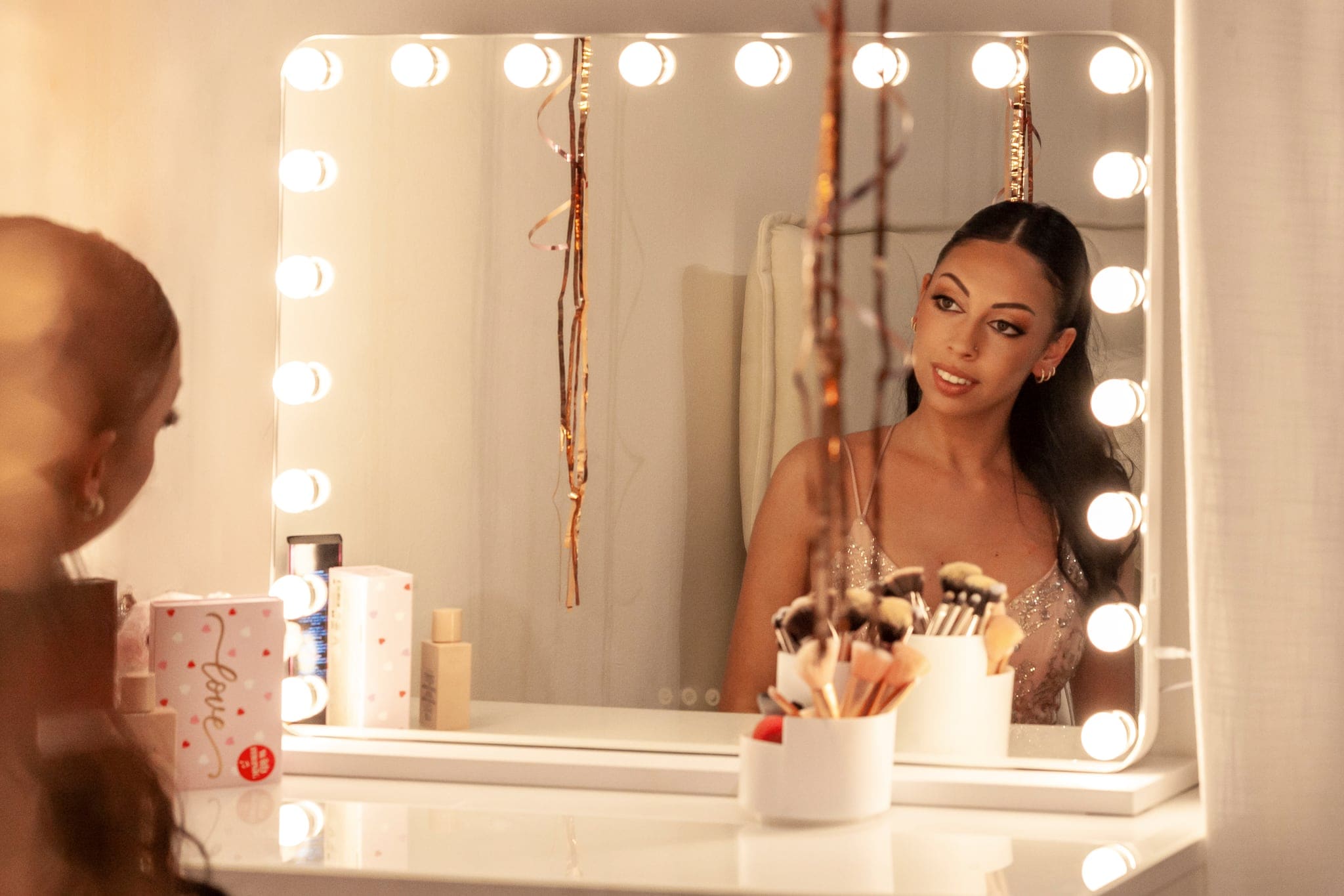 Ragazza in abito rosa glitterato si guarda allo specchio con luci da camerino durante la preparazione per il suo shooting dei 18 anni