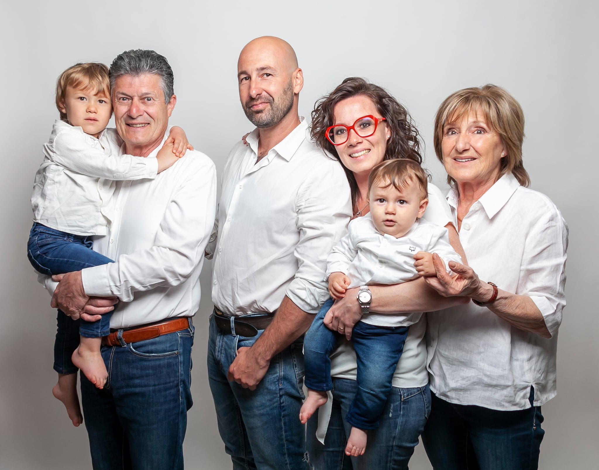 Ritratto in studio di tre generazioni in camicia bianca: nonni, genitori e due bambini piccoli in braccio, sfondo grigio chiaro