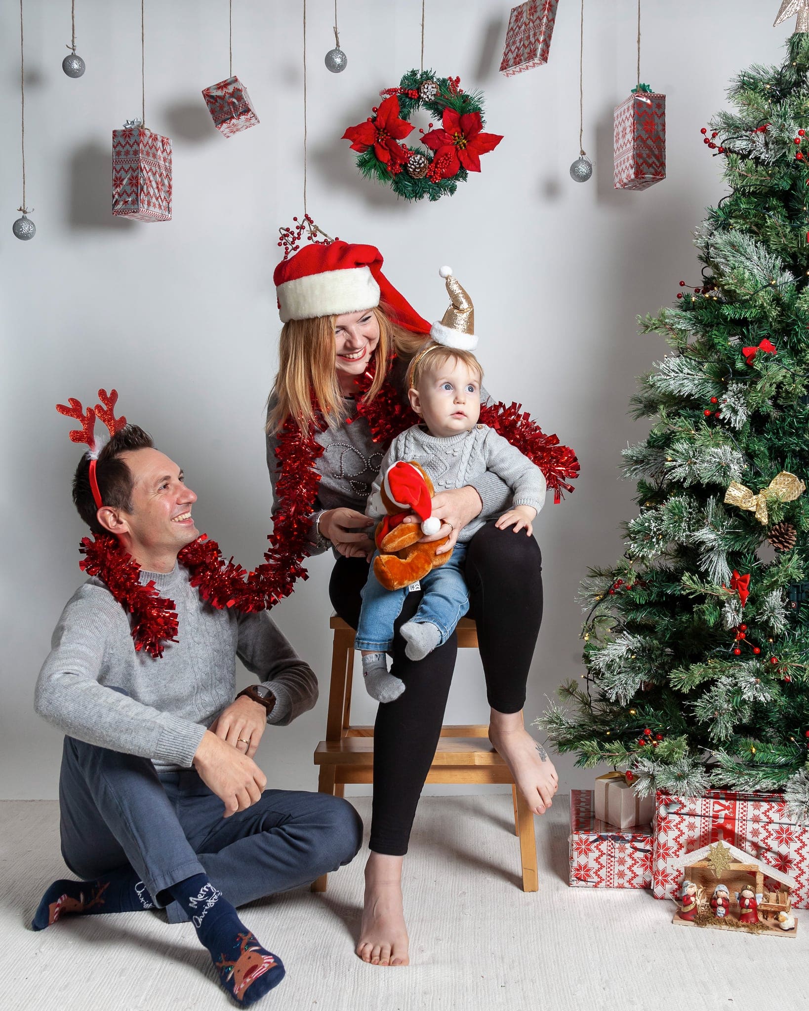 Coppia con il bambino seduto sullo sgabello, con cerchietto a renna e cappello di Babbo Natale, davanti all'albero innevato