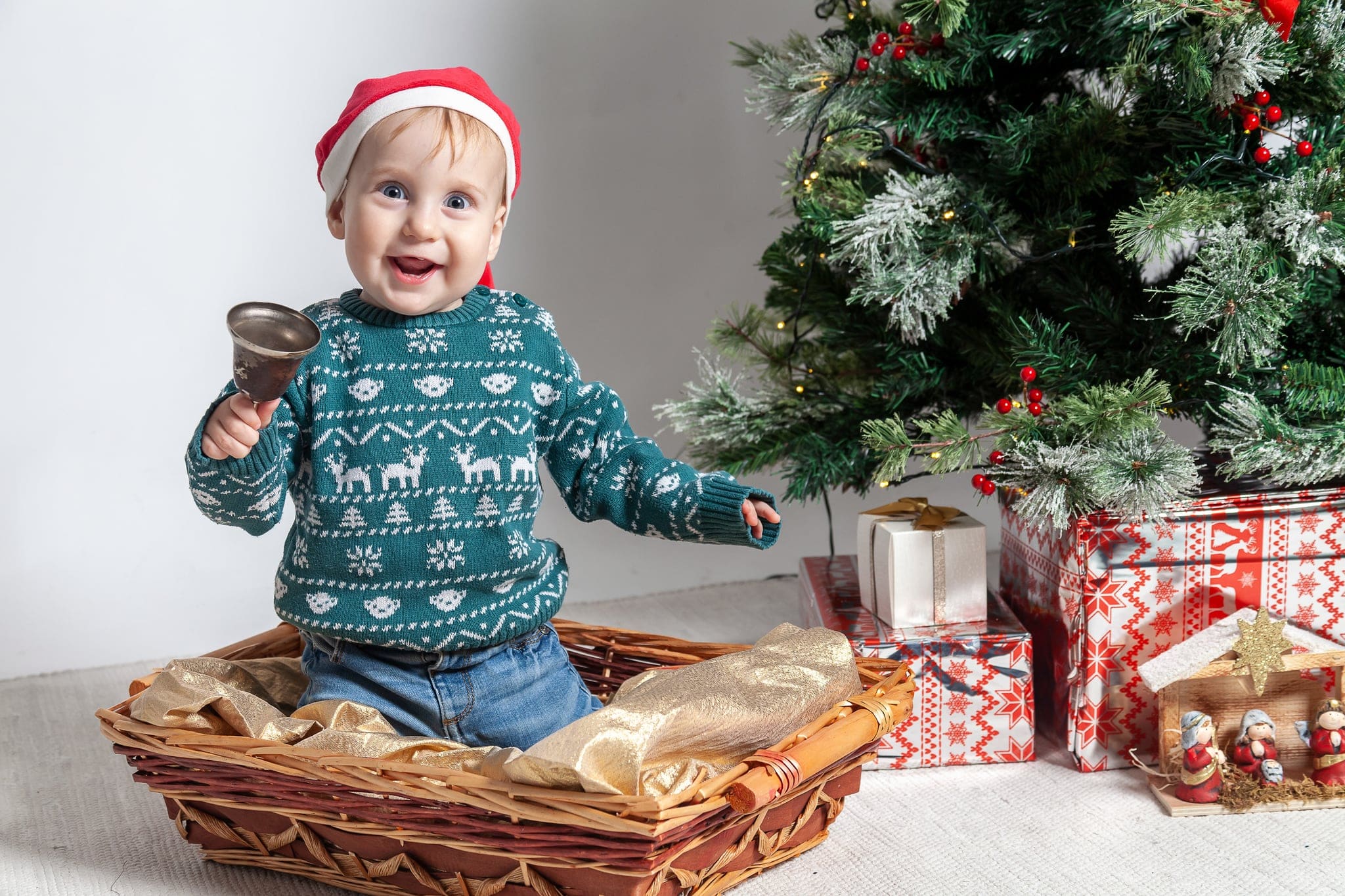 Bambino piccolo con cappellino rosso e maglione con renne ride seduto in un cesto e suona un campanello, accanto all'albero e ai regali