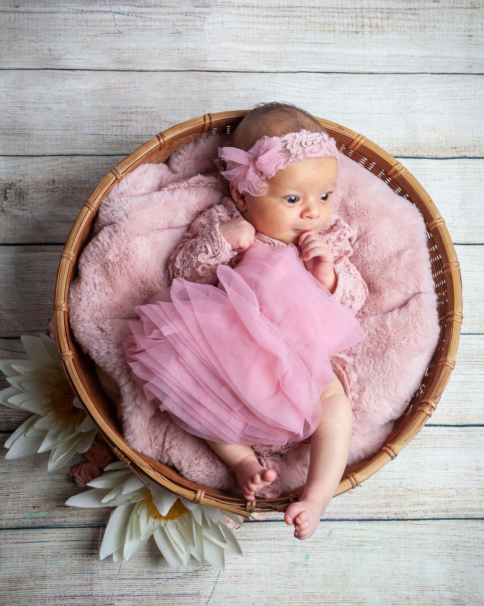 Bebè con fascia e tutù rosa sdraiata in un cesto di vimini su pelo rosa, con ninfee bianche e assi di legno chiaro.
