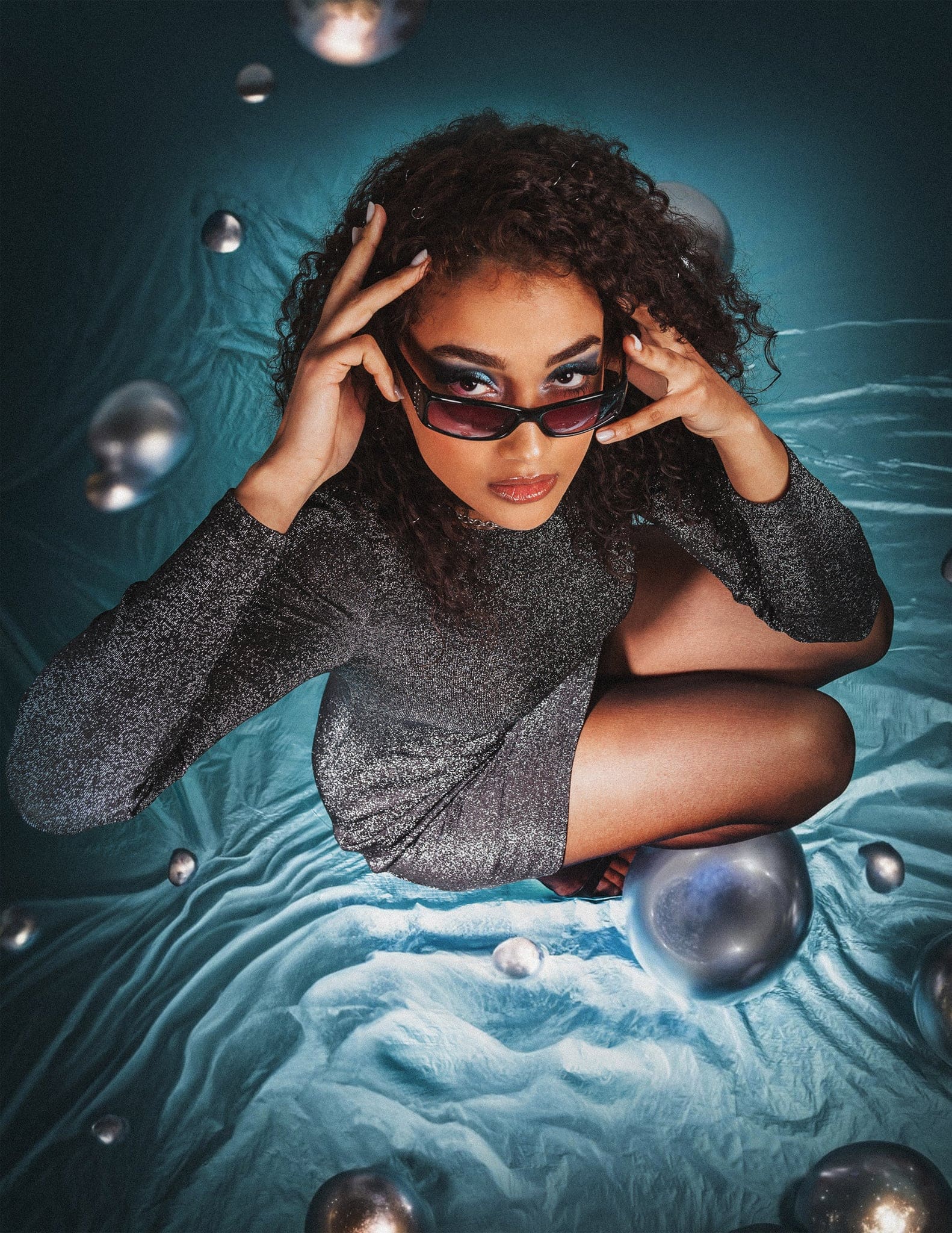 Ragazza dai capelli ricci con occhiali scuri e maglia luccicante, accovacciata su un telo blu con sfere argentate, in stile creativo