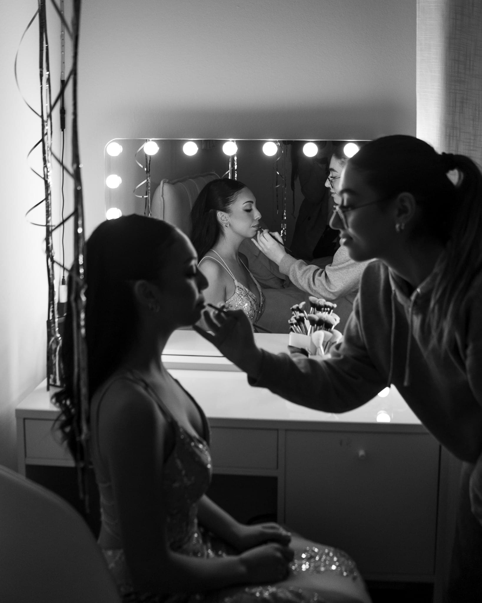 In bianco e nero, la truccatrice sistema il make-up alla festeggiata seduta davanti allo specchio con le luci, prima della festa dei 18 anni