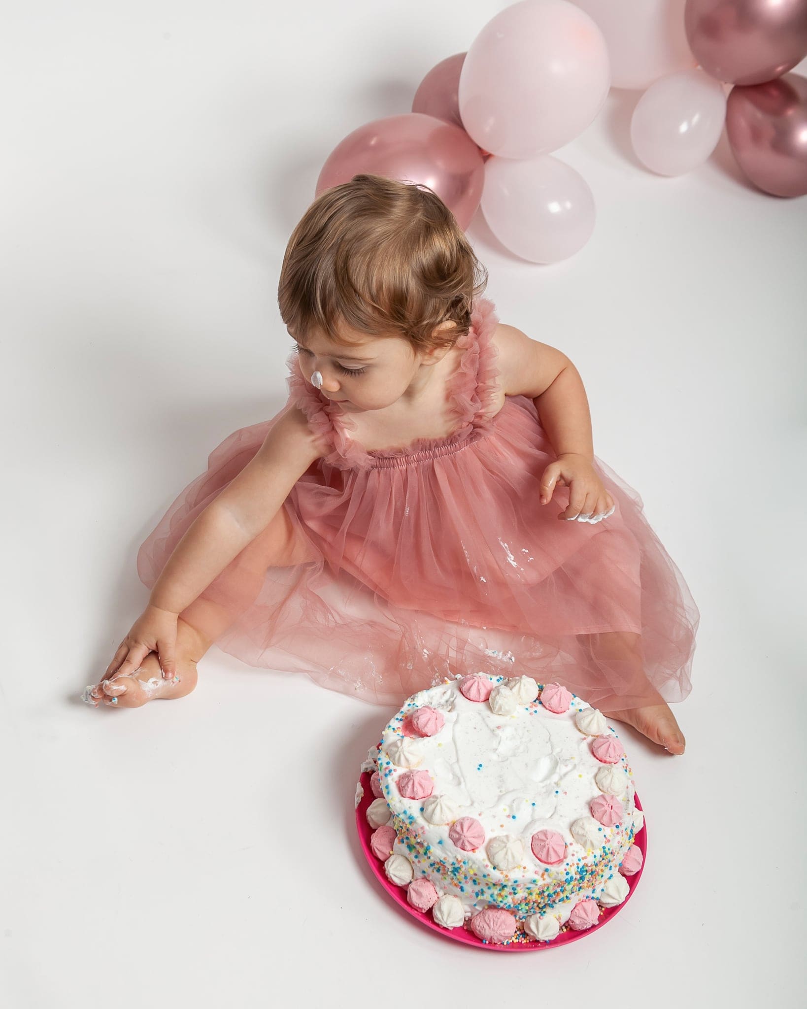 Dall'alto, la piccola in tulle rosa siede sul fondale bianco con panna sul naso e sulle mani, accanto alla torta