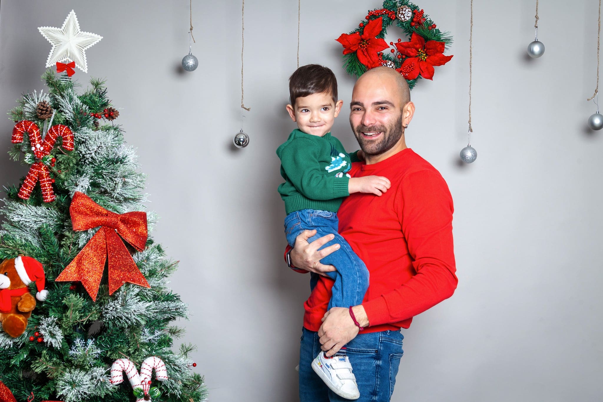 Papà con maglione rosso sorride tenendo in braccio il figlio in maglione verde, davanti all'albero di Natale e alla ghirlanda