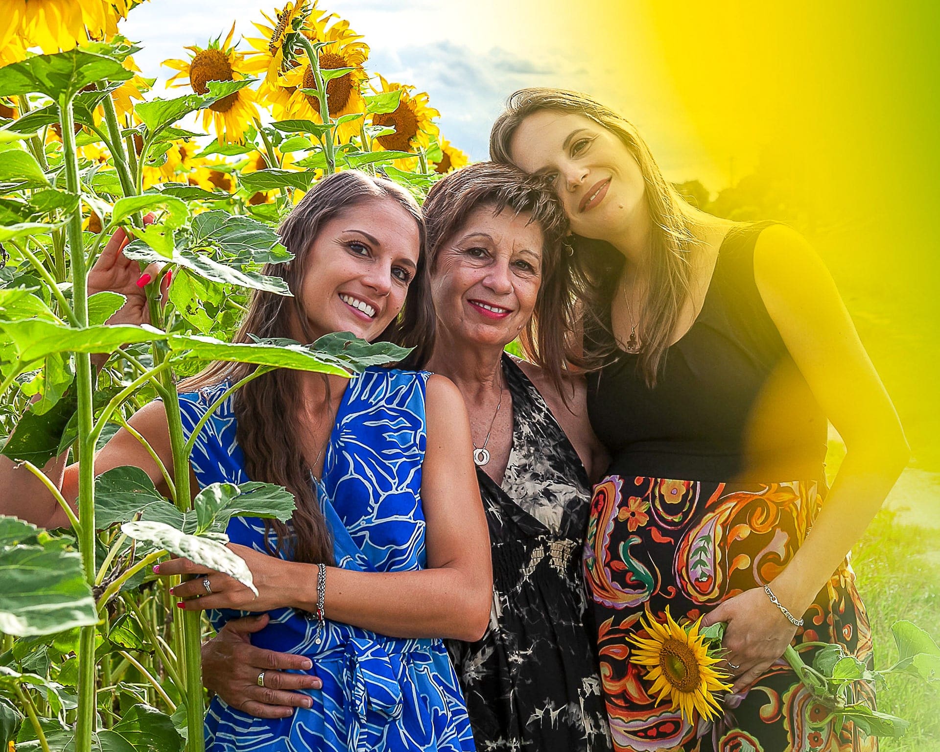 Tre donne di età diverse abbracciate tra i girasoli, con luce dorata e una di loro in dolce attesa