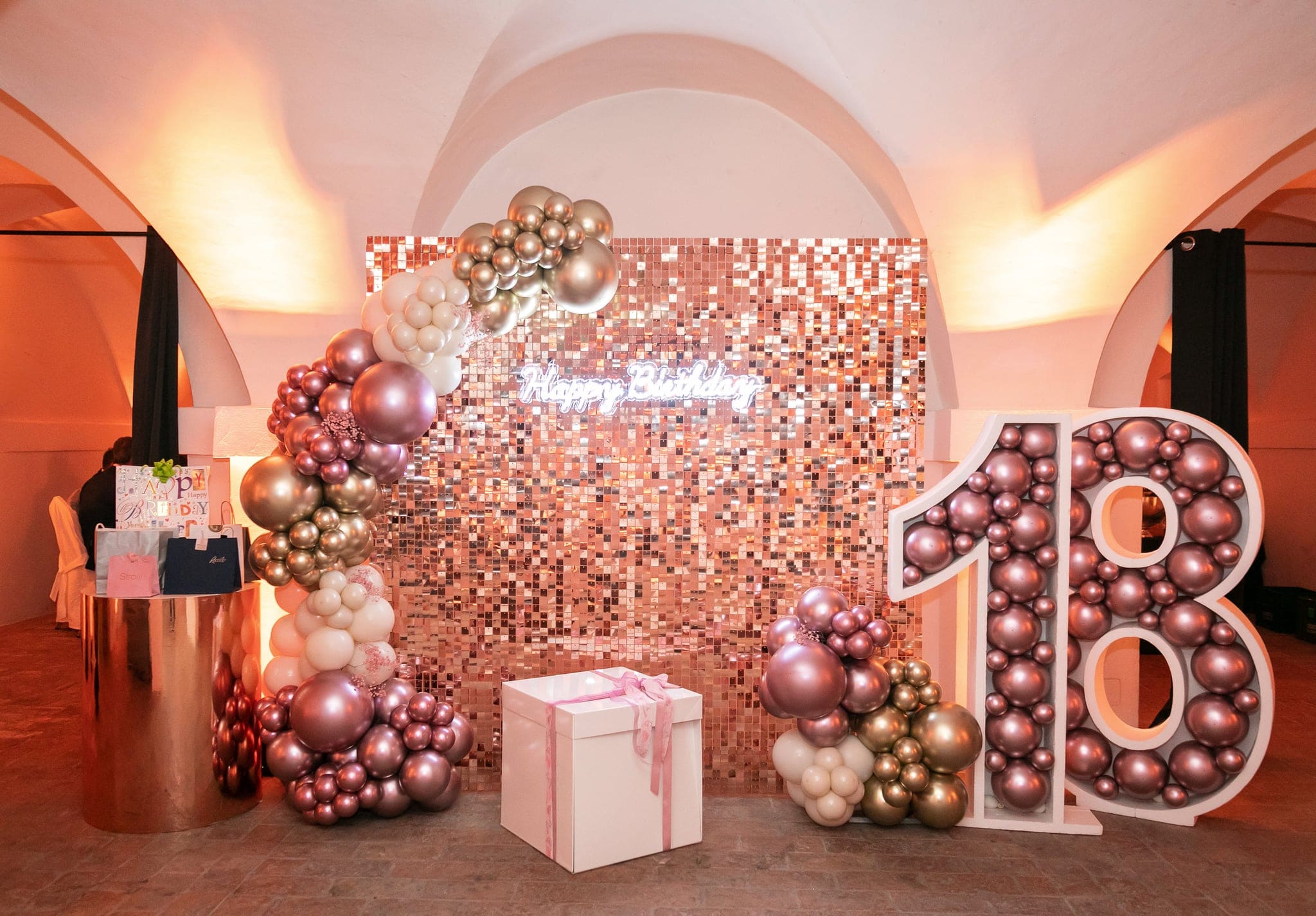 Allestimento per i 18 anni con parete di paillettes rosa, scritta Happy Birthday, grande numero 18 di palloncini e regalo con fiocco, in una sala a volte