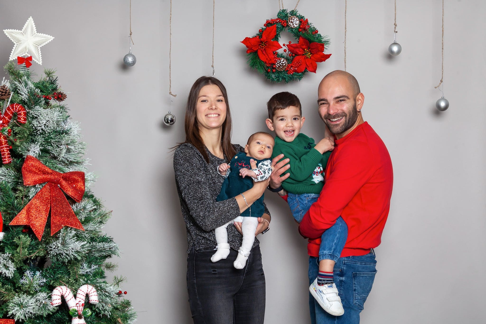 Famiglia in posa sullo sfondo grigio: mamma con la neonata, papà con il bimbo in braccio, accanto all'albero e alla ghirlanda