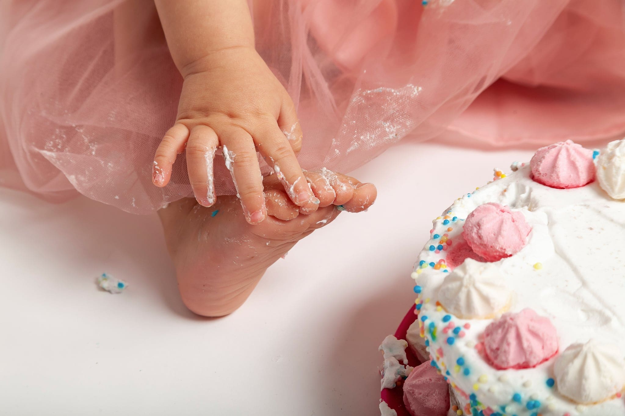 Dettaglio della manina sporca di panna che stringe il piedino, accanto a una torta con meringhe rosa e codette colorate