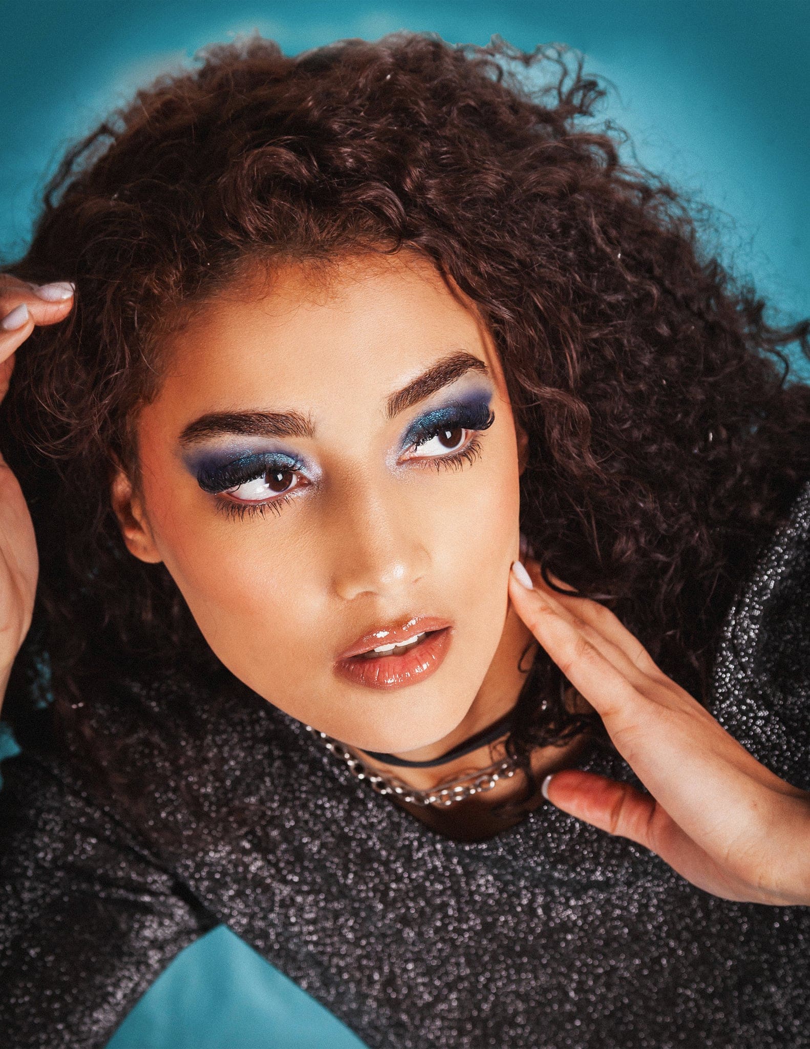 Primo piano di una ragazza dai folti ricci con trucco blu e brillante, mano sul viso, su fondo turchese