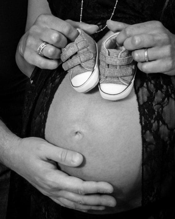 Scarpine da neonato tenute dalla mamma sopra il pancione, con la mano del papà che lo accarezza, in bianco e nero.
