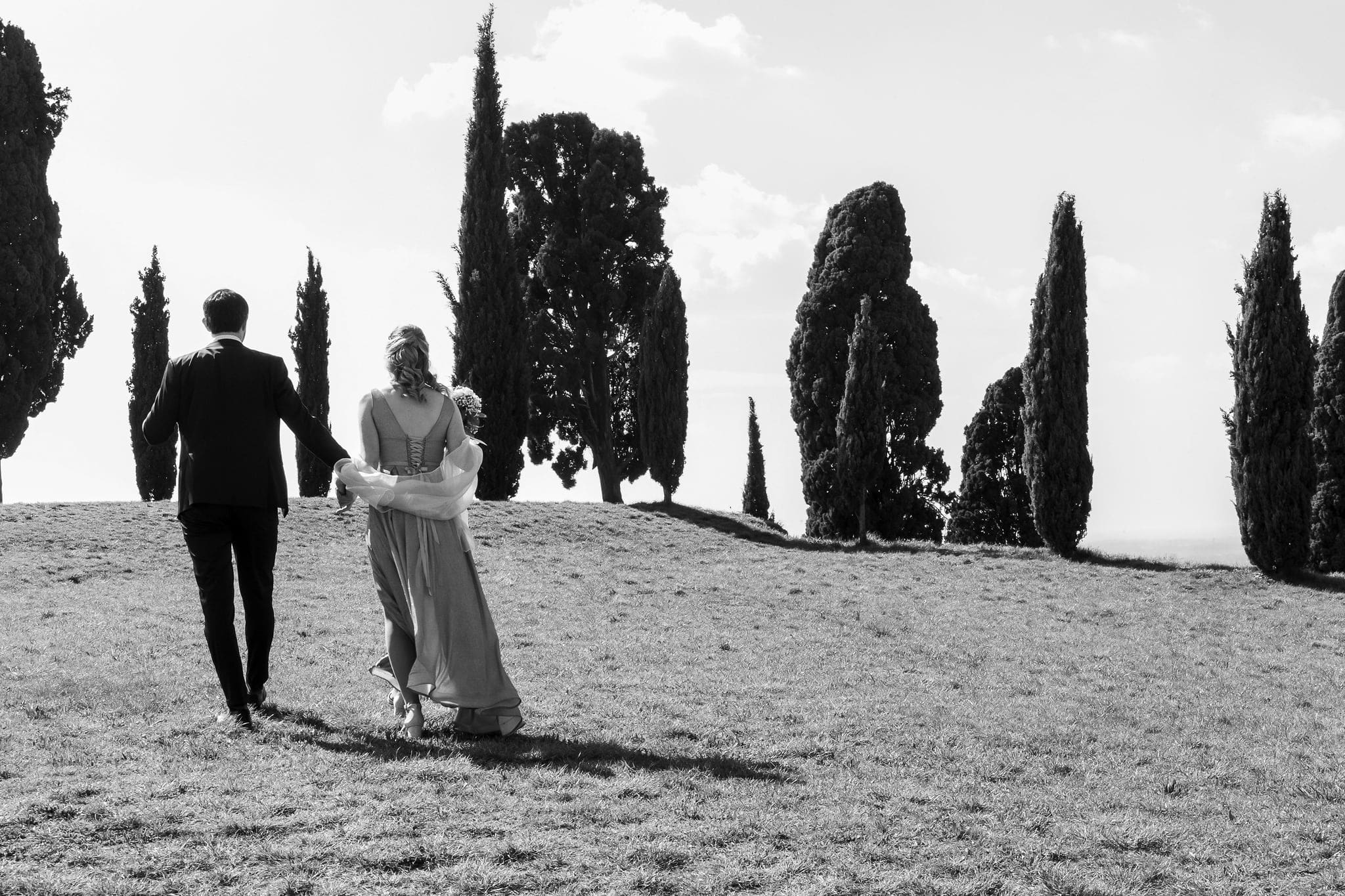 Sposi che camminano mano nella mano su un prato verso i cipressi, in bianco e nero, matrimonio a Fagagna