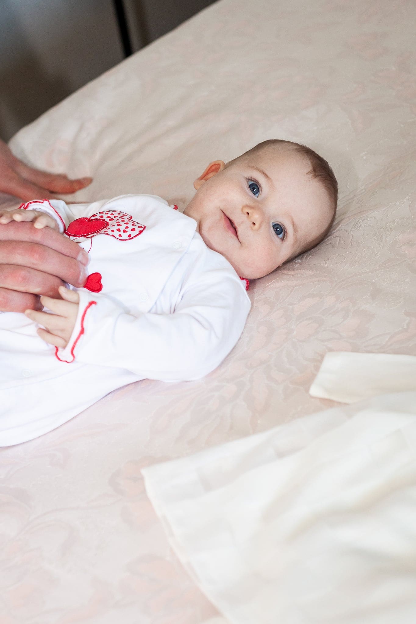 Una bimba sorridente con gli occhi azzurri è sdraiata sul letto in tutina bianca, accanto al vestitino del battesimo