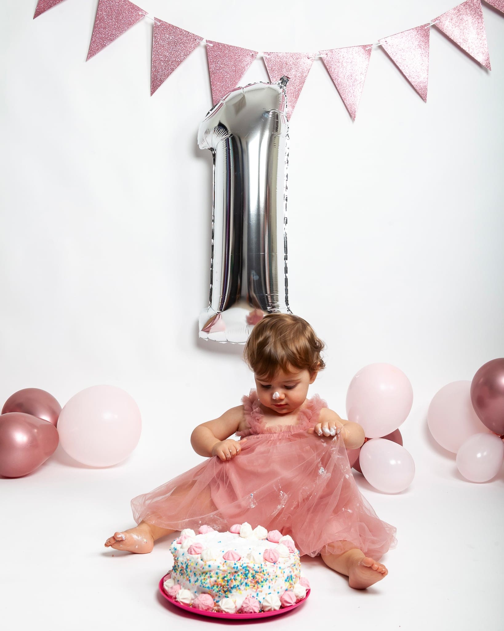 Smash cake in studio: la bimba in abito rosa osserva la torta, con festone rosa e palloncino argentato a forma di uno