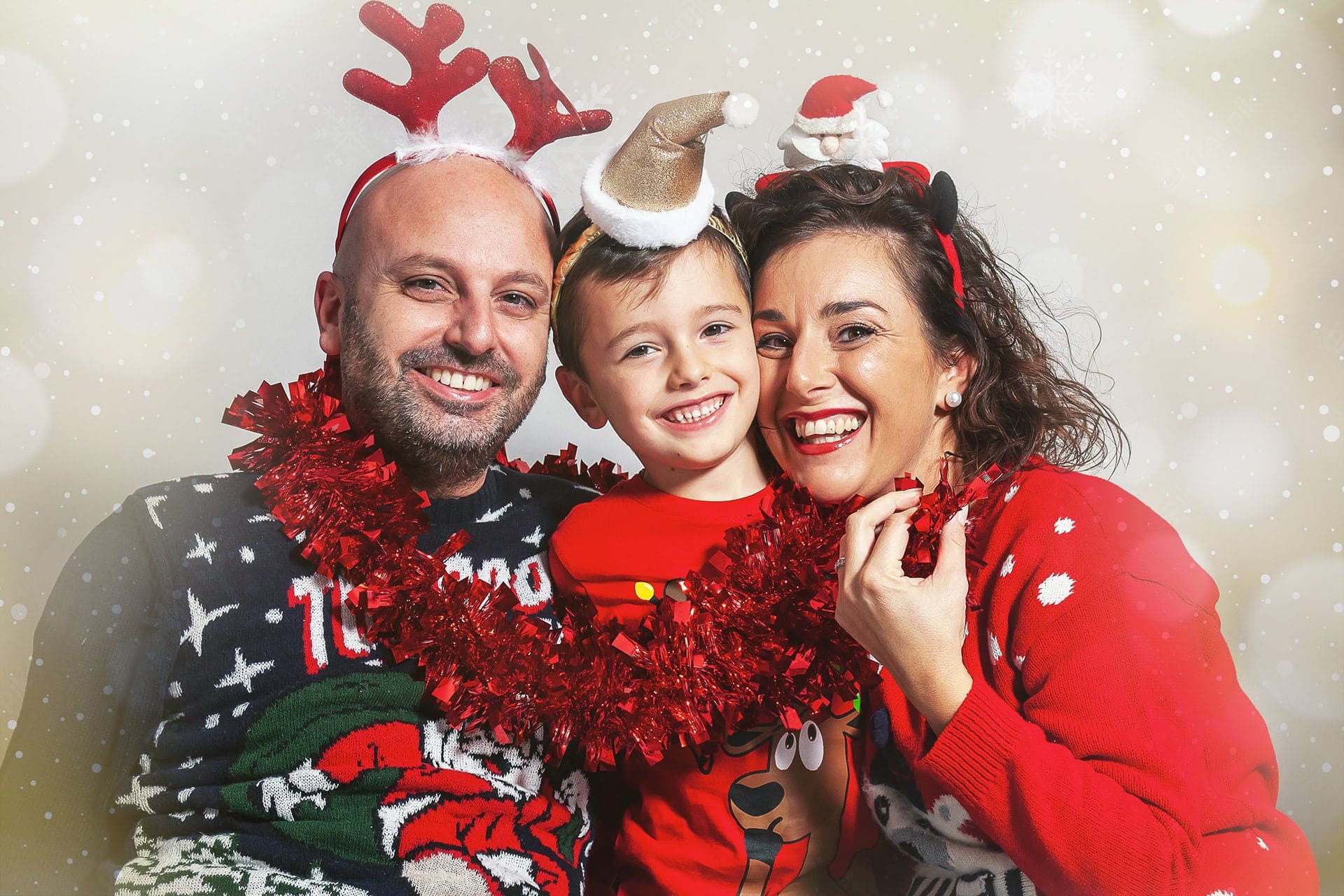 Primo piano di mamma, papà e figlio che ridono vicini, con maglioni natalizi, tinsel rosso e cerchietti, su fondo con luci e neve