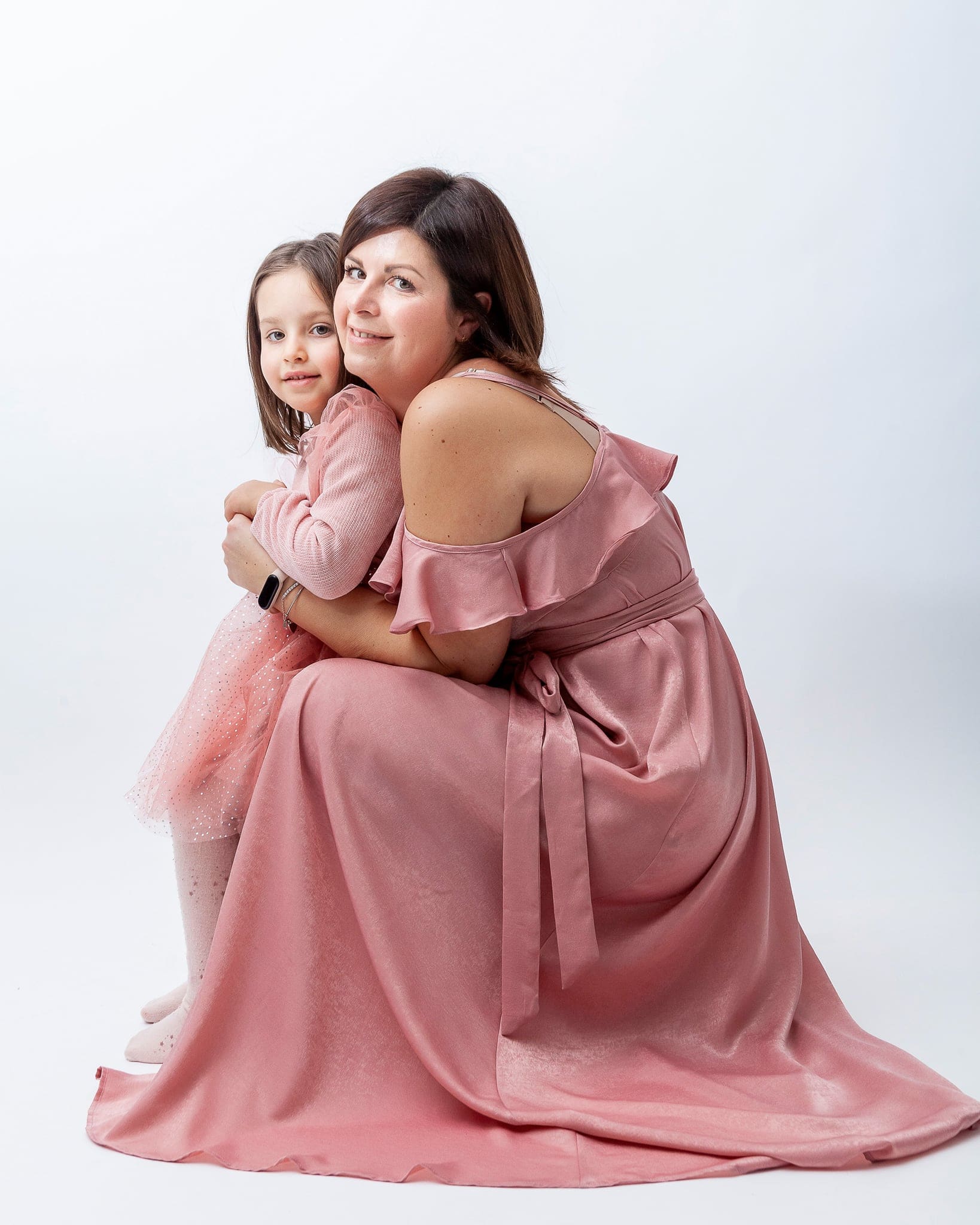 Mamma in abito rosa lungo seduta sui talloni abbraccia la figlia in tulle rosa, su sfondo bianco in studio