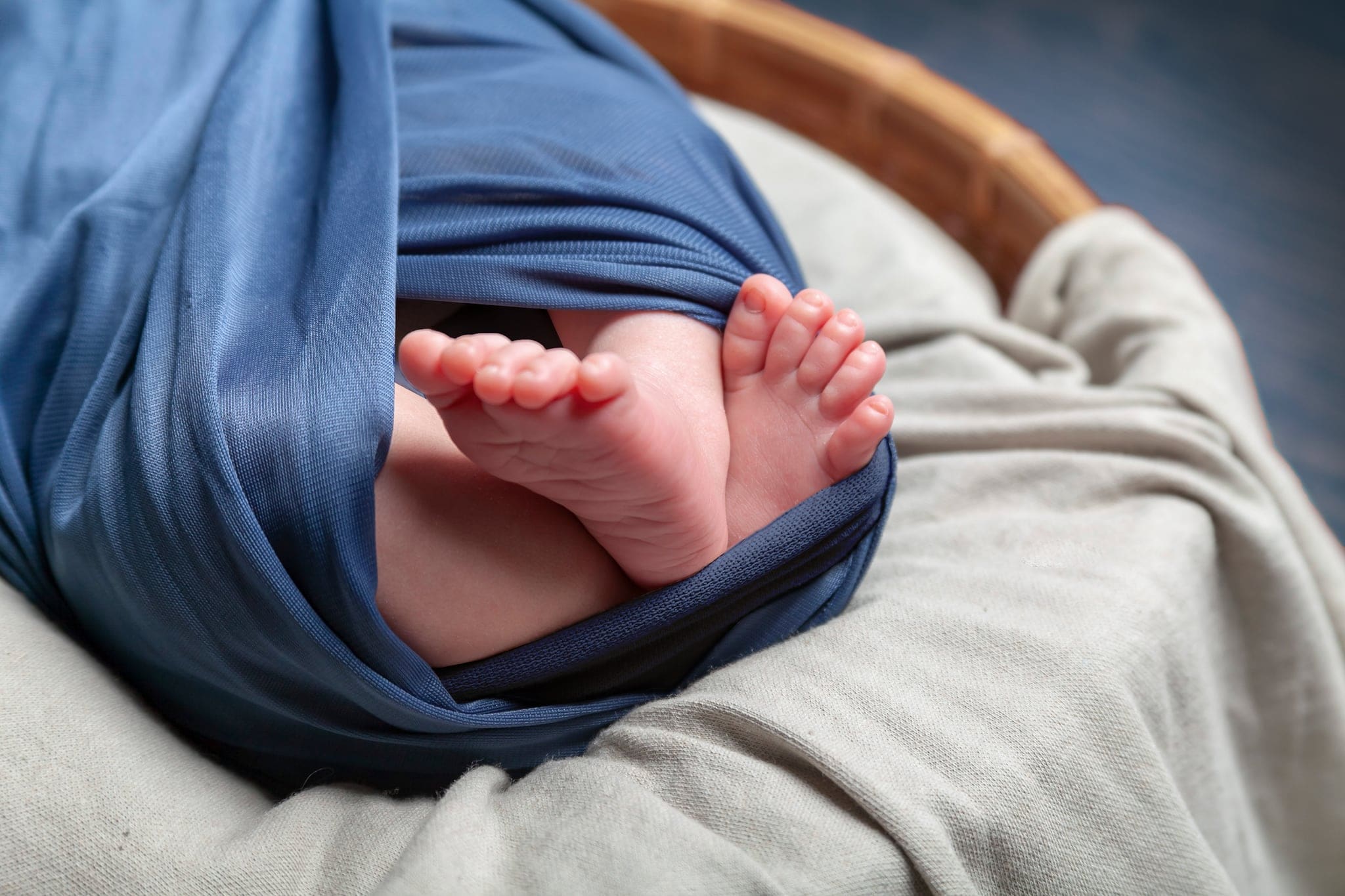 Piedini di un neonato che sbucano da un telo blu avvolto, in un cesto con coperta beige, in un servizio newborn in studio.