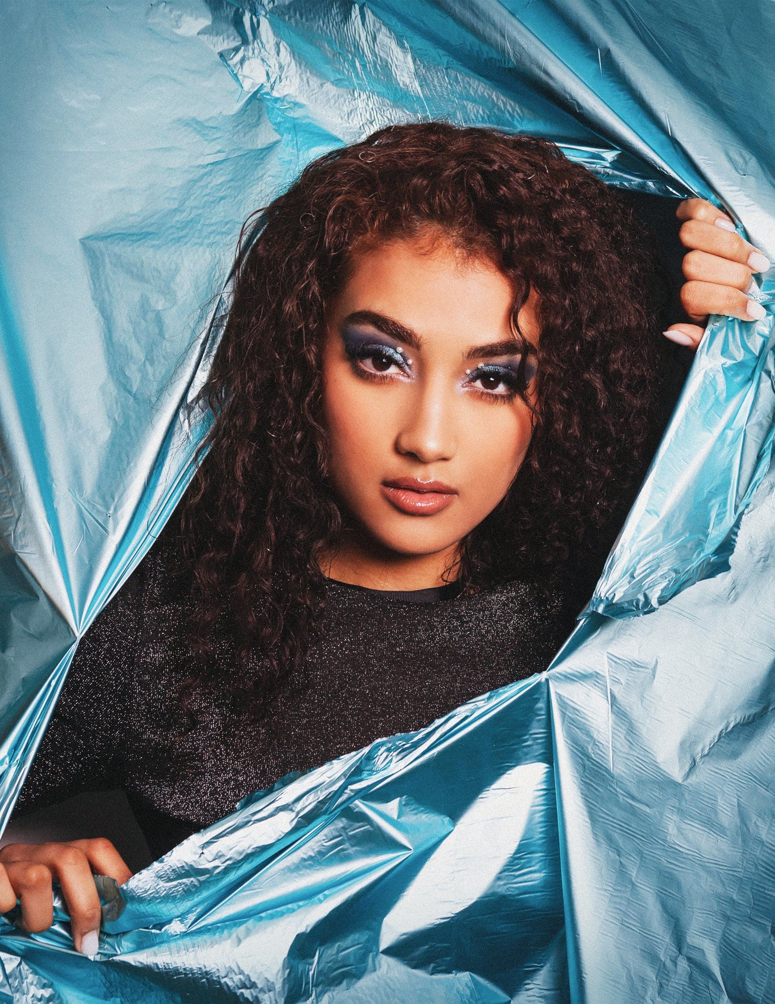 Ragazza dai capelli ricci con trucco blu guarda in camera attraverso uno squarcio in un foglio metallizzato azzurro