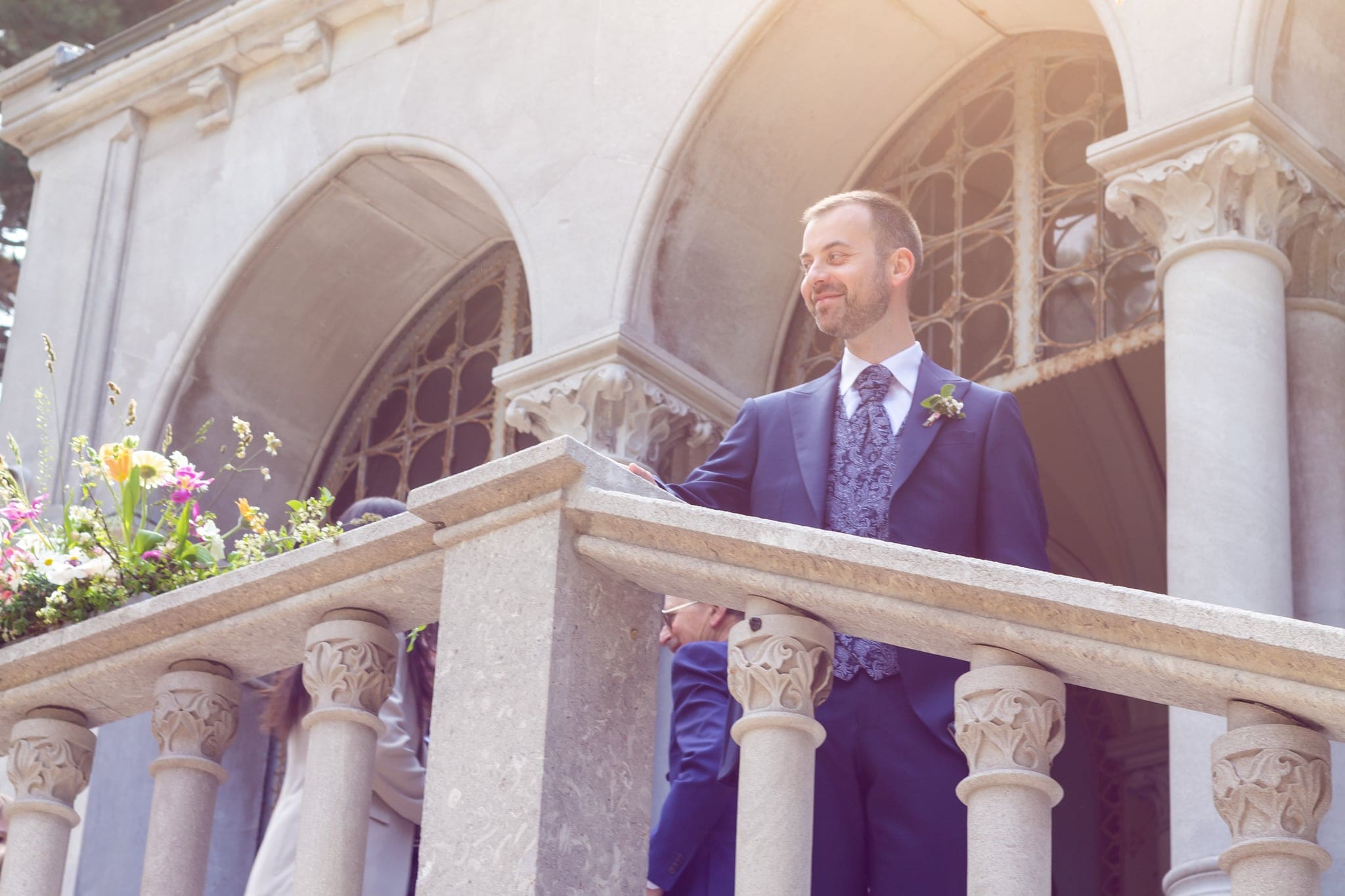 Sposo sorridente affacciato a un balcone con colonne in pietra e fiori, in attesa della sposa, matrimonio a Trieste