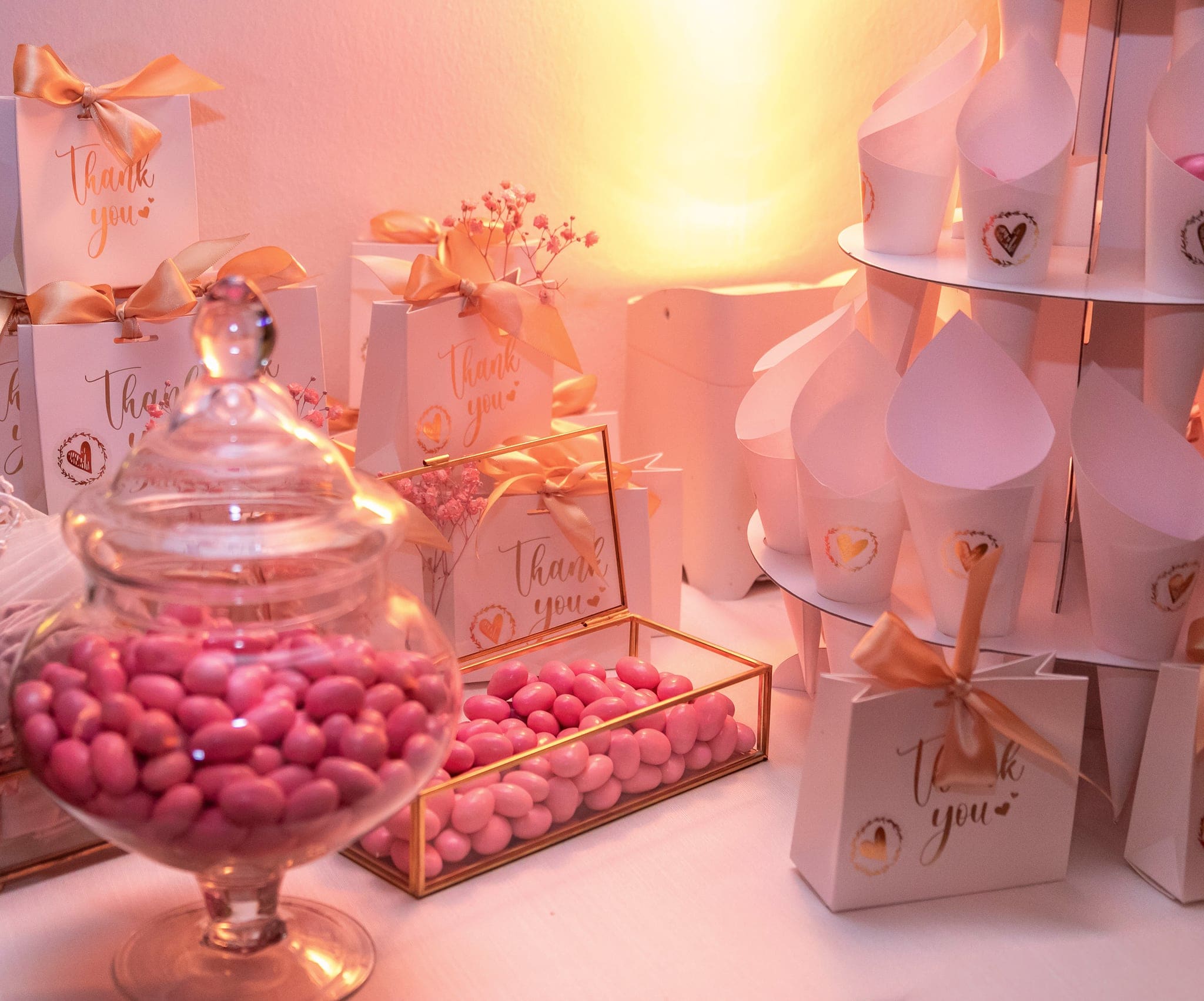 Angolo dei dolci con vasi di vetro pieni di confetti rosa e sacchettini Thank you con fiocchi dorati, in luce calda