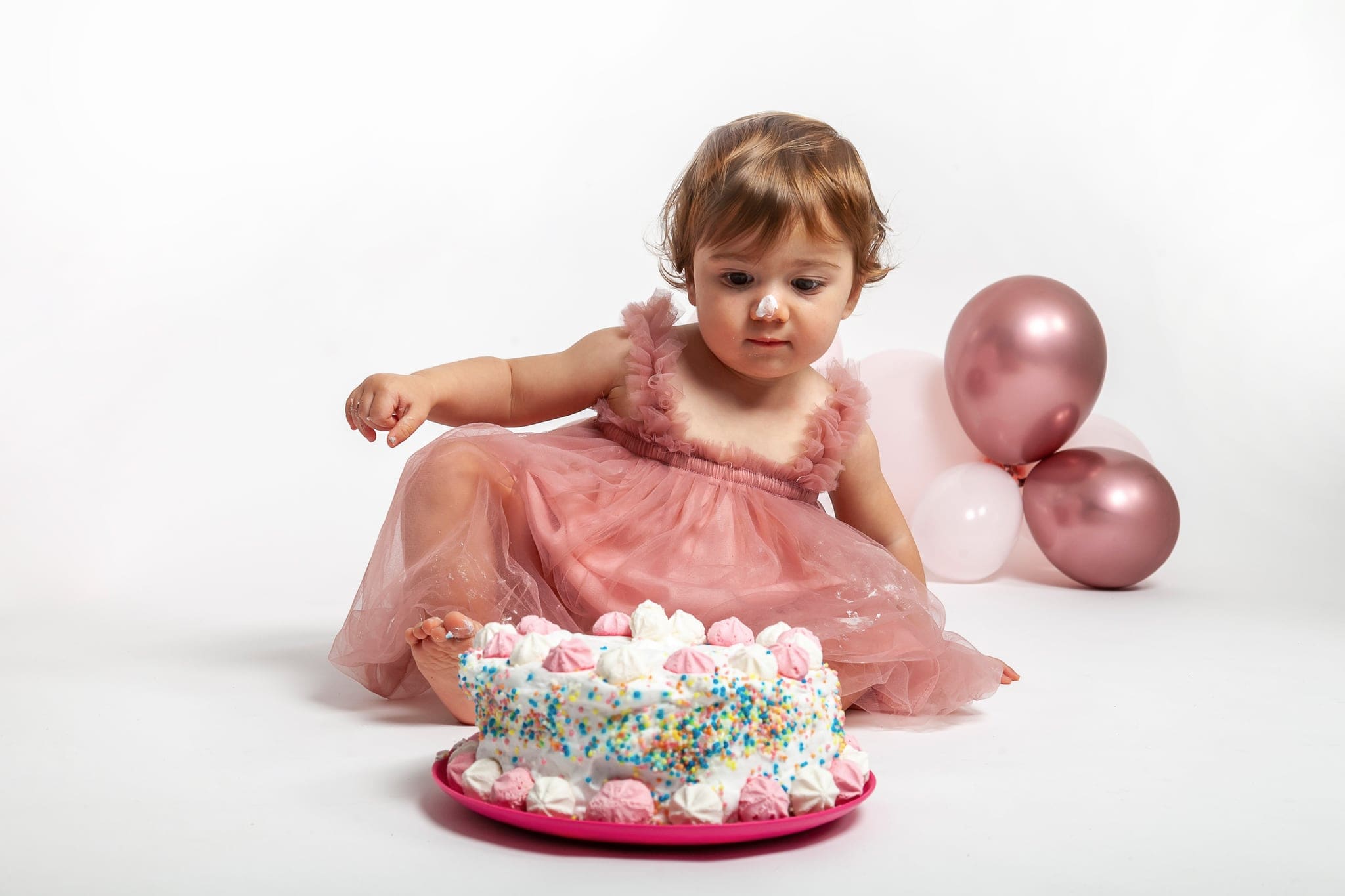 Seduta davanti alla torta con le meringhe rosa, la piccola allunga una mano mentre guarda con curiosità, vicino ai palloncini rosa