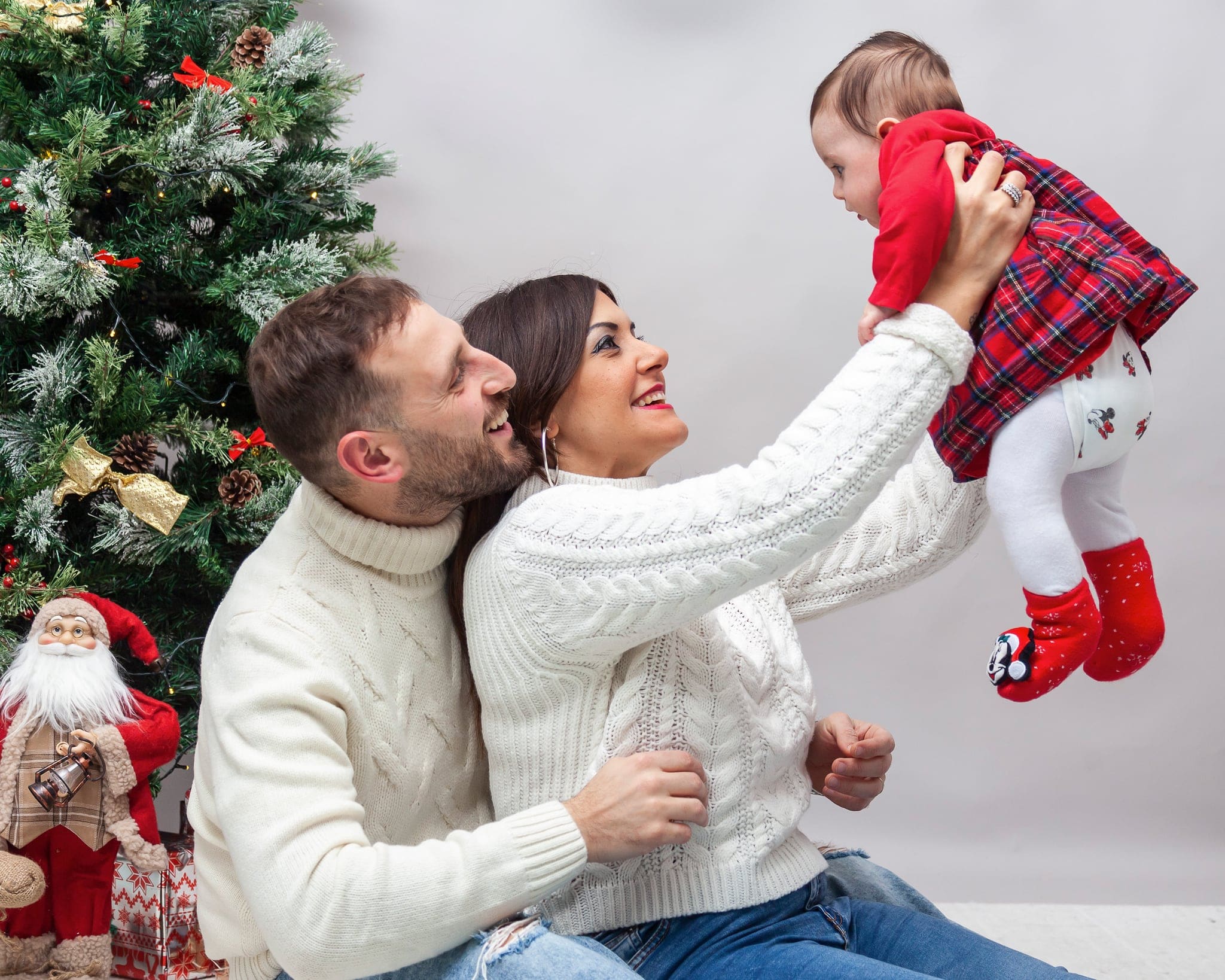 Coppia in maglioni bianchi sorride sollevando la bimba con vestitino a quadri rosso, accanto all'albero e a Babbo Natale