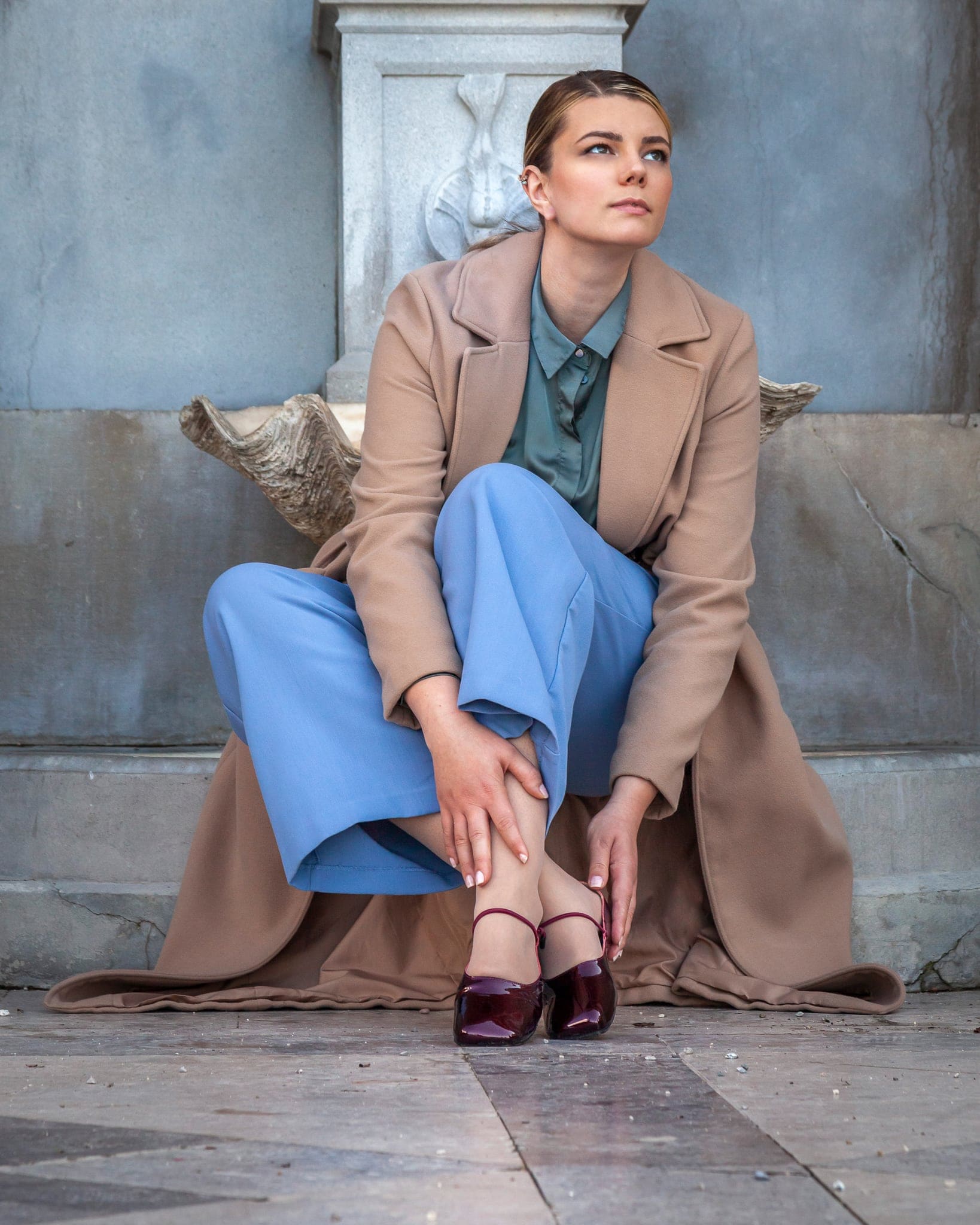Donna in cappotto beige e pantaloni azzurri accovacciata davanti a un muro in pietra con elemento scolpito, sguardo verso l'alto