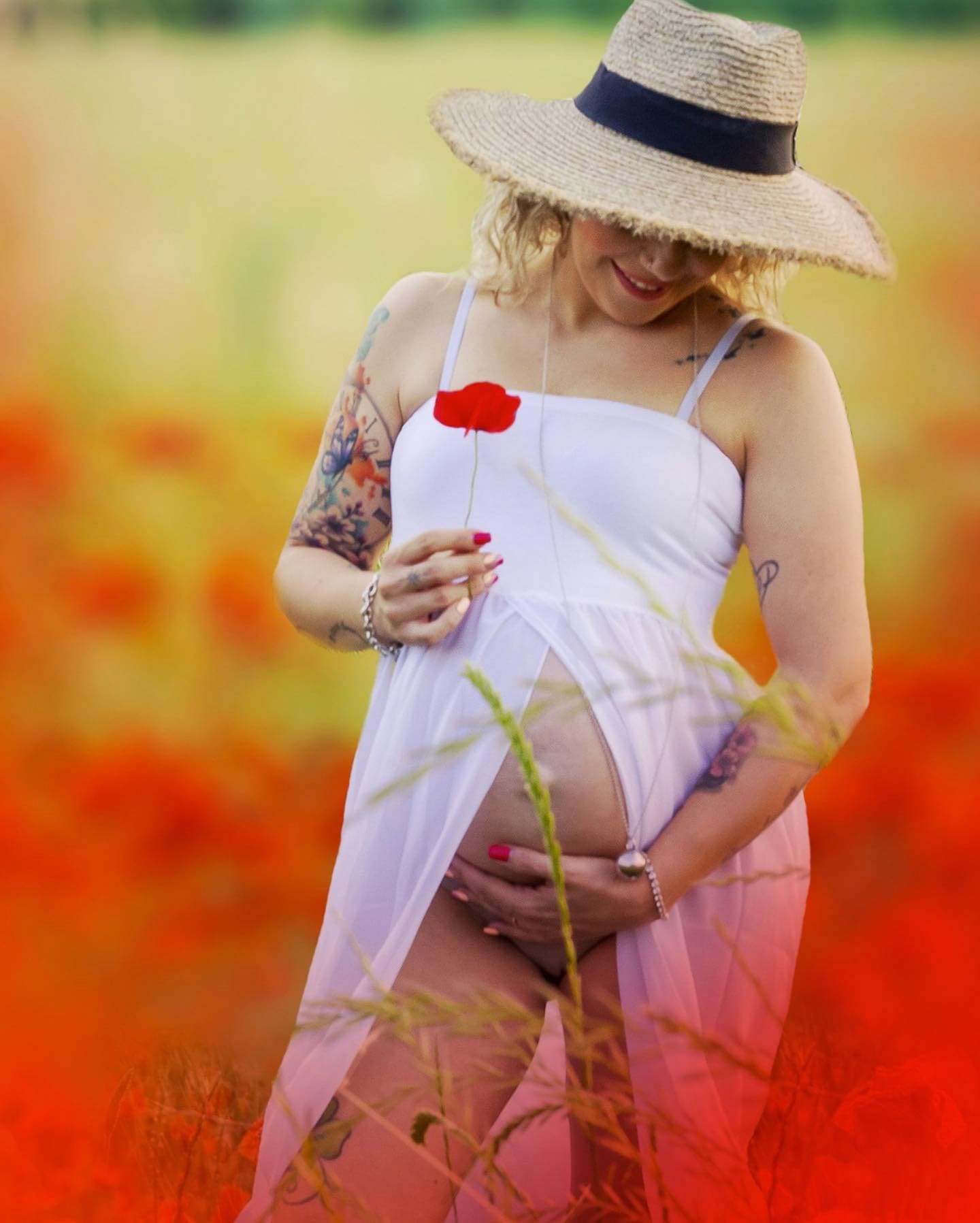 Donna incinta in un campo di papaveri con cappello di paglia e abito bianco, regge un papavero e tocca il pancione.