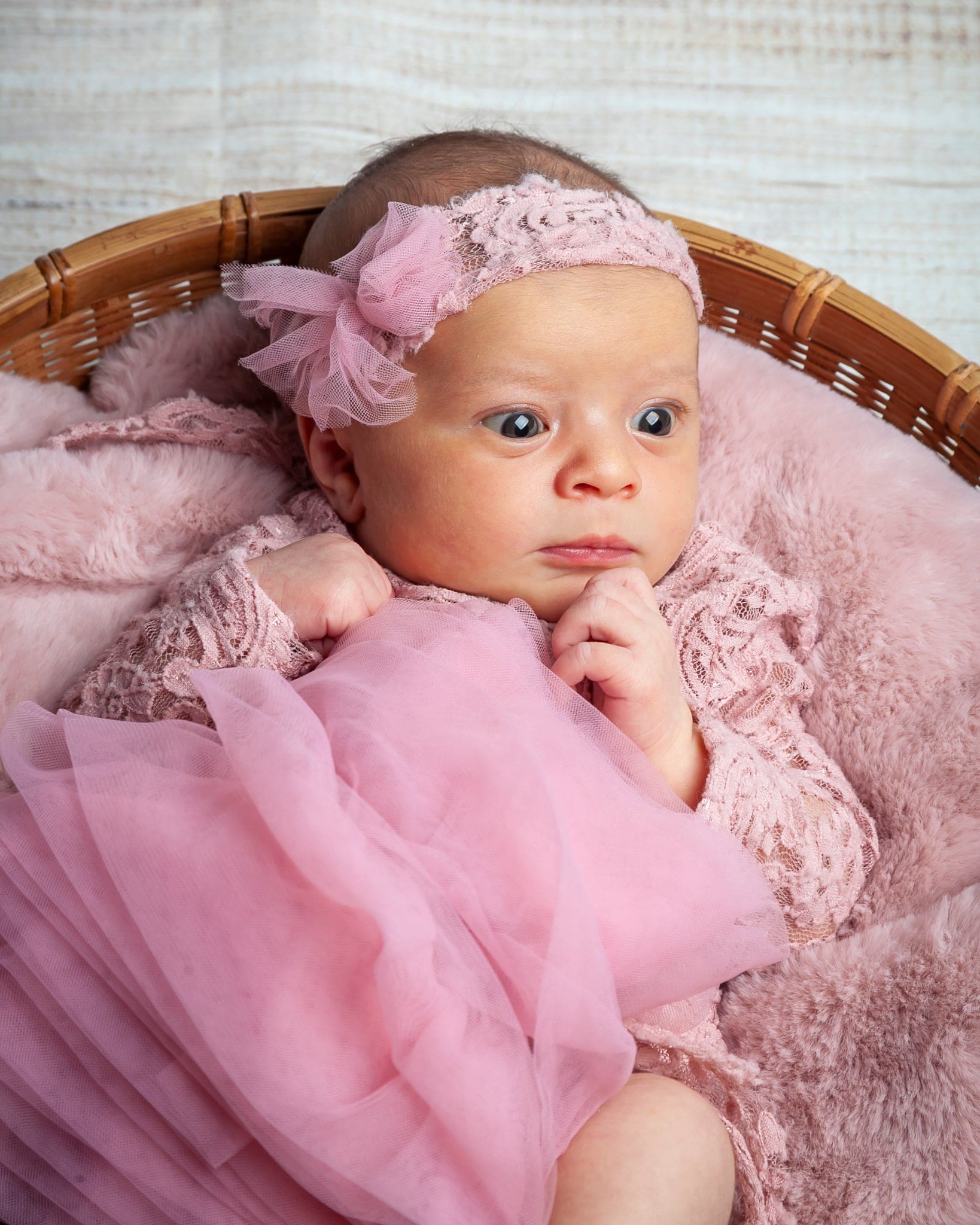 Primo piano di una neonata con fascia a fiocco, body di pizzo e tulle rosa, con la mano sul mento e lo sguardo attento.
