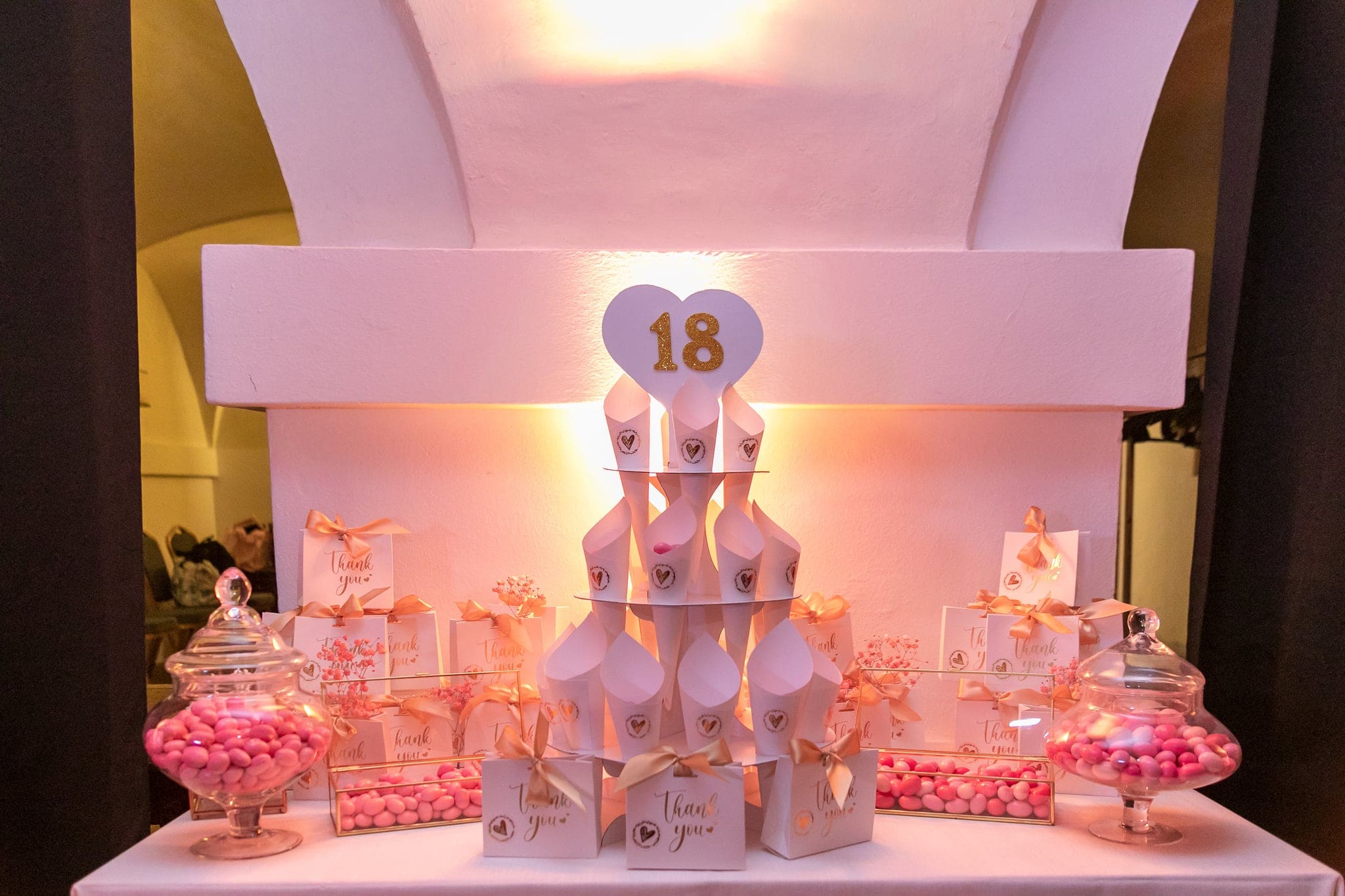 Tavolo dei confetti con torre di coni bianchi, cuore con il numero 18 e vasi di confetti rosa, nella sala a volte illuminata di arancio