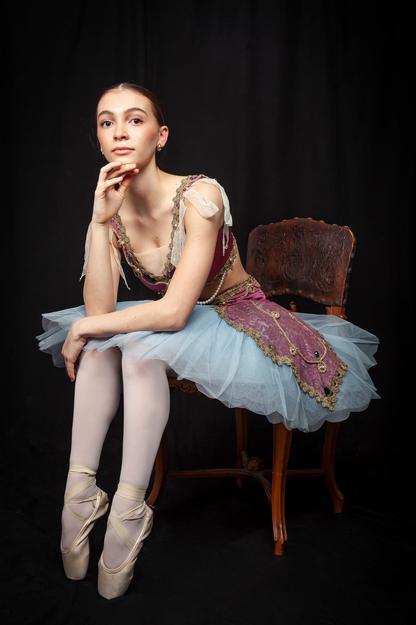 Giovane ballerina in tutù azzurro e corpetto viola e oro, in scarpe a punta, seduta su una sedia antica con la mano al mento su fondo nero