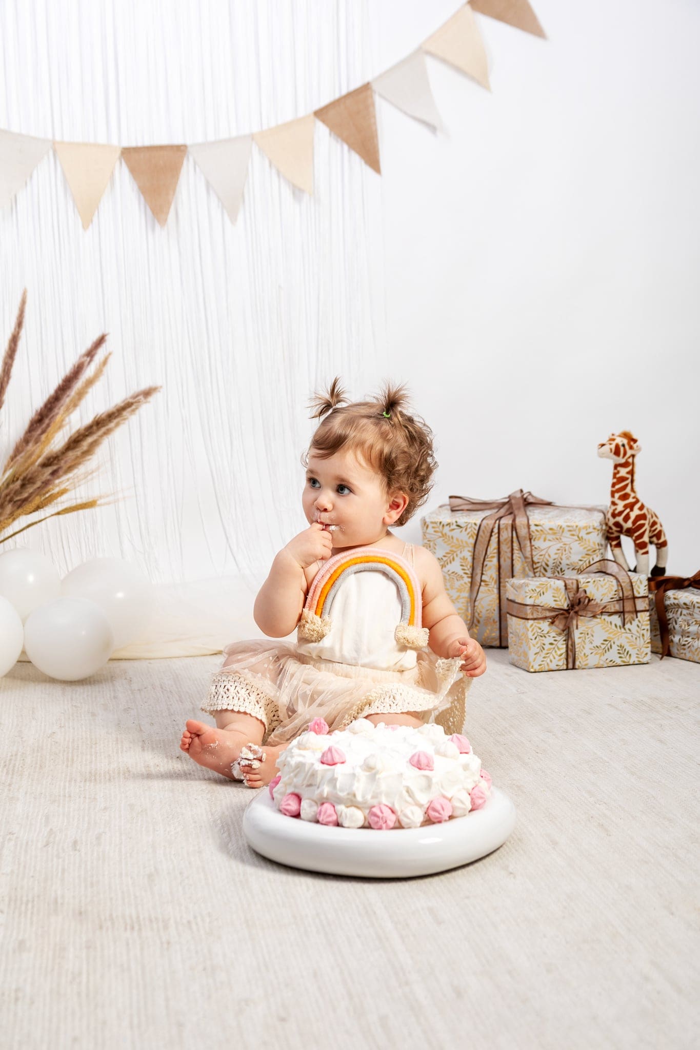Allestimento boho con festone beige e fiori secchi: la piccola, con un arcobaleno di lana sulla tutina, si porta la mano alla bocca