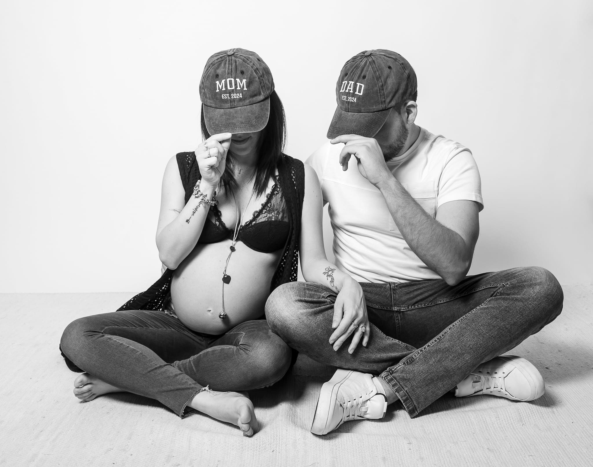 Coppia in attesa seduta a gambe incrociate con cappellini con scritto MOM e DAD che nasconde il volto, in bianco e nero.