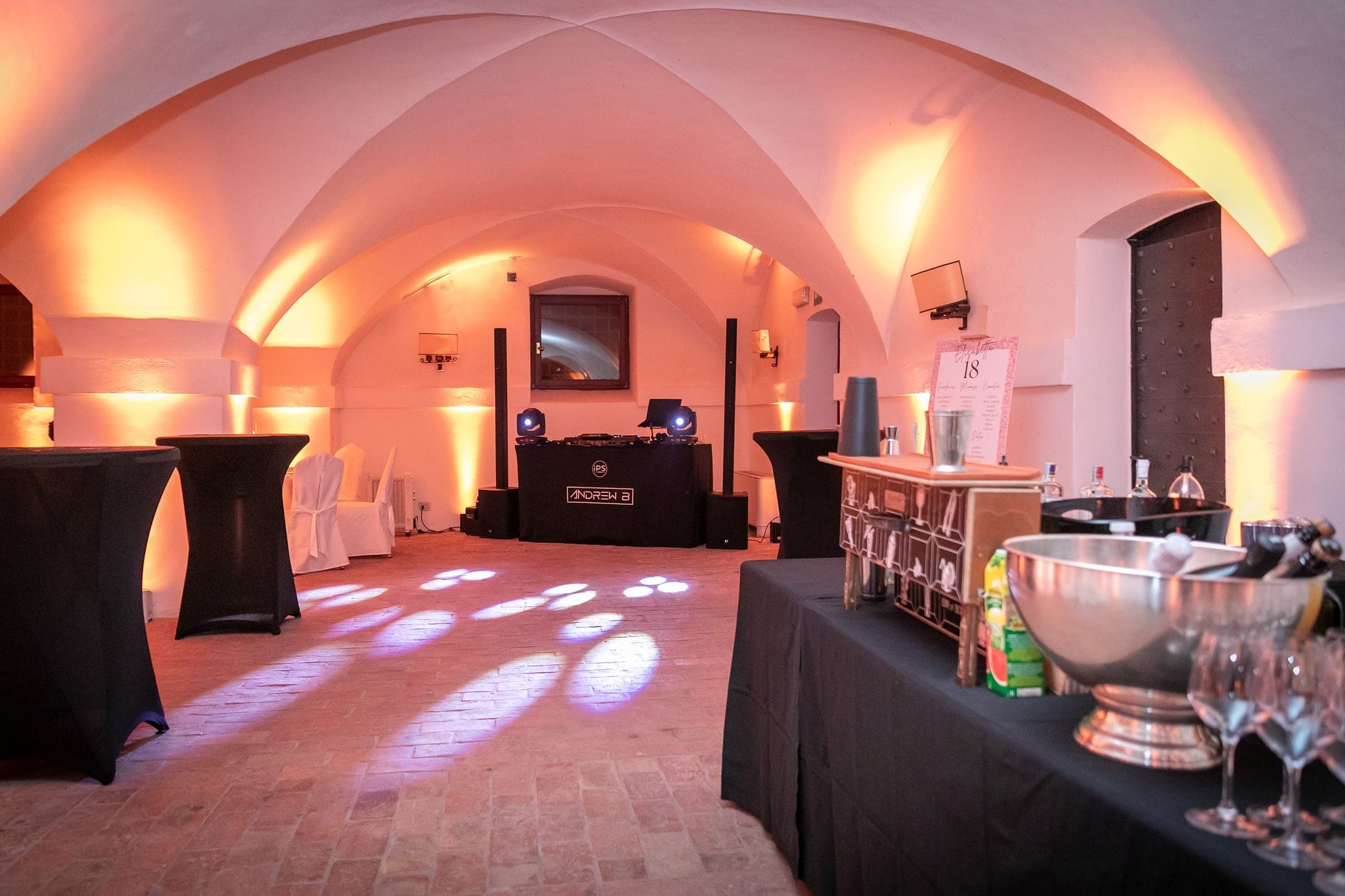 Sala con soffitto a volte illuminata da luci calde, tavolini alti neri, consolle del dj e angolo bar pronti per la festa di compleanno