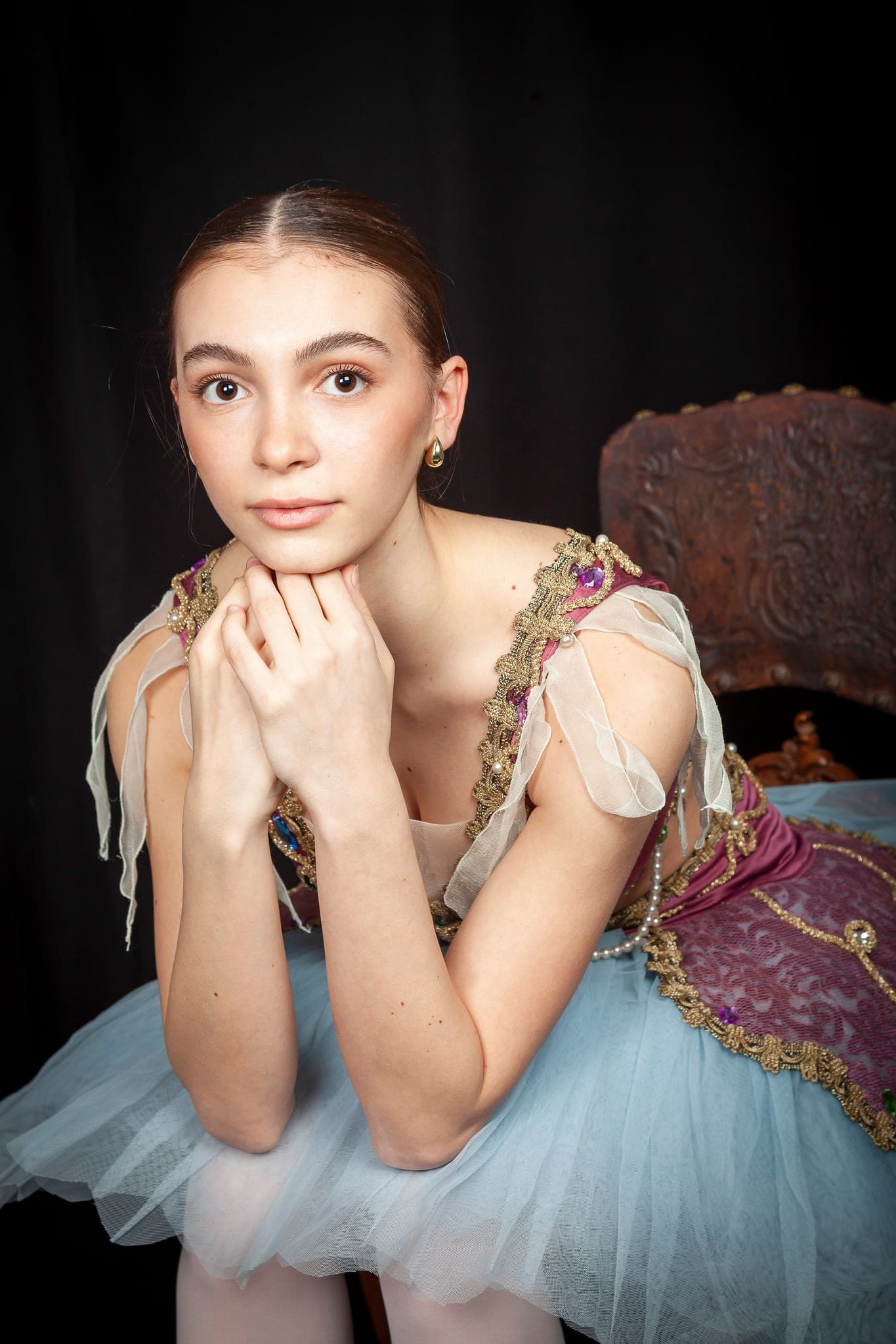 Primo piano di una giovane ballerina in costume di scena con le mani intrecciate sotto il mento, sguardo dolce e fondo scuro