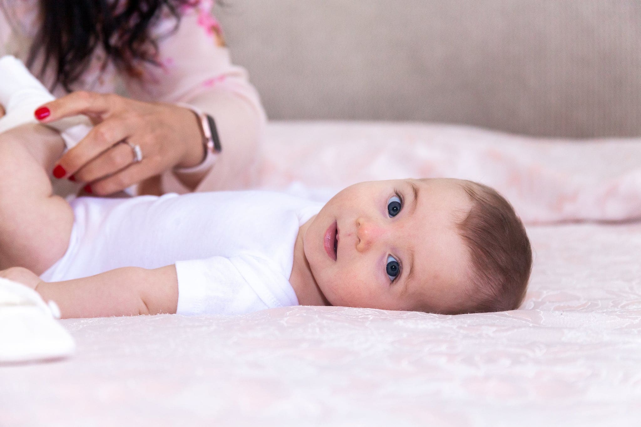 La piccola è sdraiata sul letto con lo sguardo curioso, mentre la mano della mamma le tiene il piedino