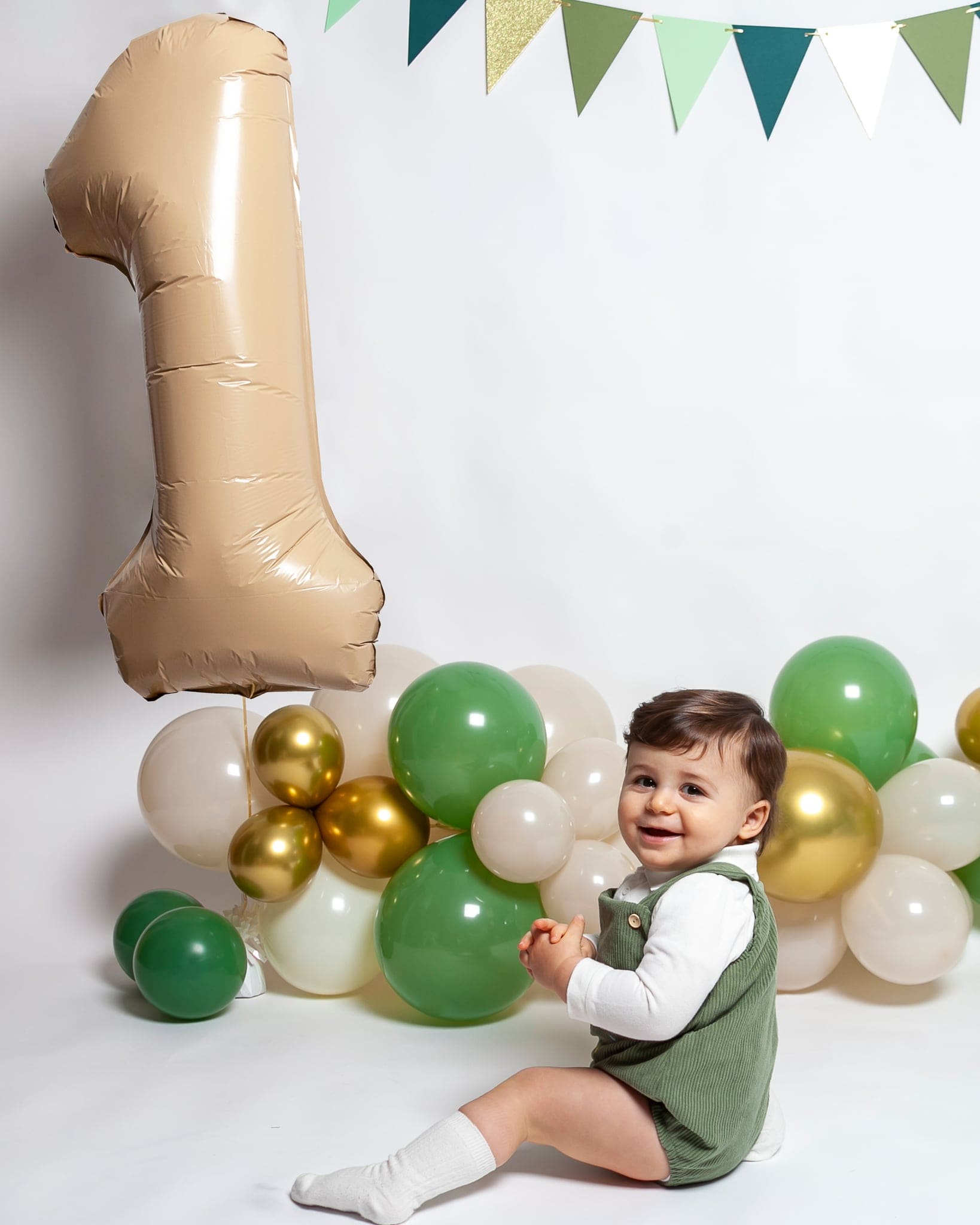 Un bimbo sorridente in salopette verde siede accanto a una ghirlanda di palloncini verdi, oro e beige e a un grande uno color sabbia