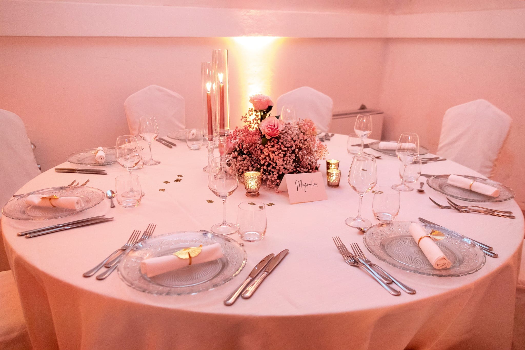 Tavolo rotondo apparecchiato con piatti, bicchieri, candele e centrotavola di fiori rosa, nella luce rosata della sala