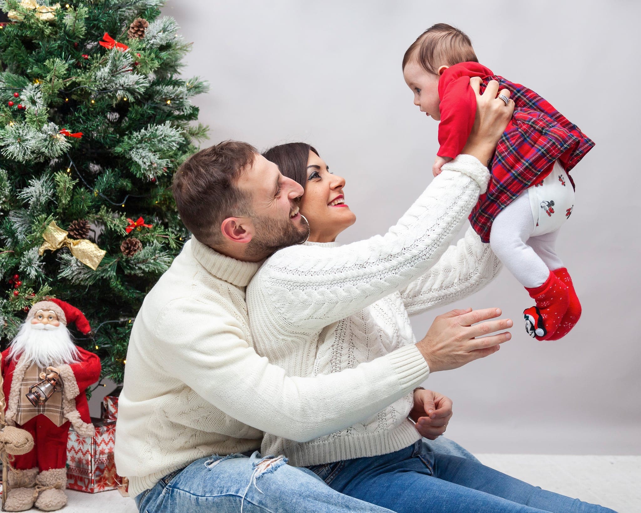 Mamma e papà in maglioni bianchi sollevano in aria la figlia in abito rosso, ridendo, accanto all'albero di Natale
