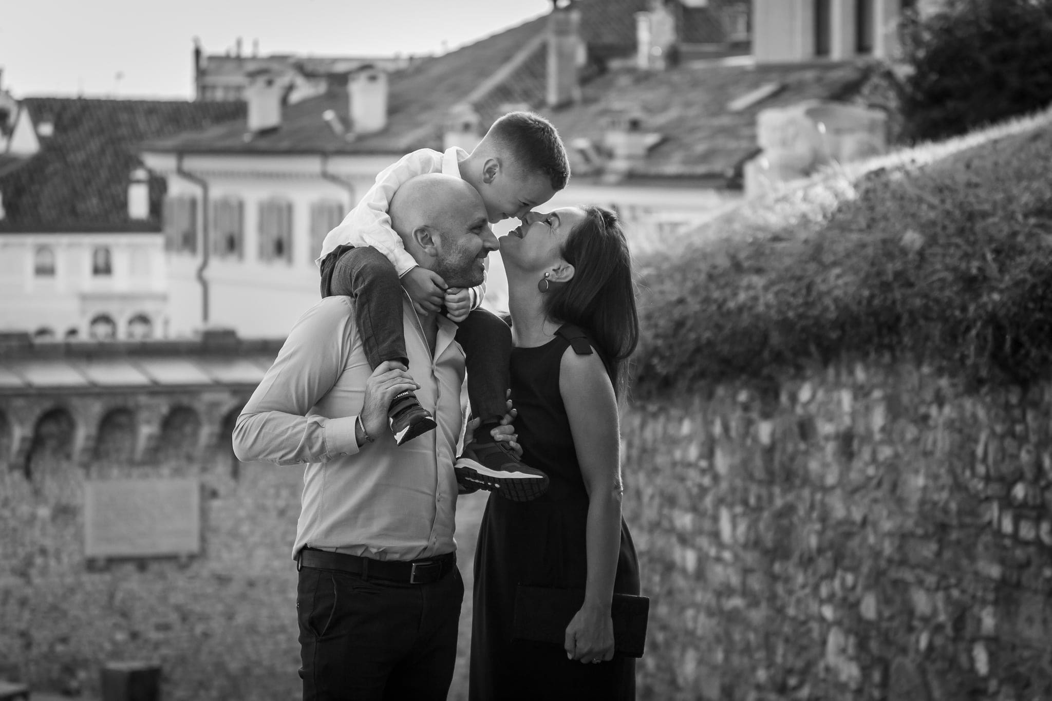 Papà con il bambino sulle spalle bacia la mamma davanti a un muro di pietra con tetti in città, in bianco e nero
