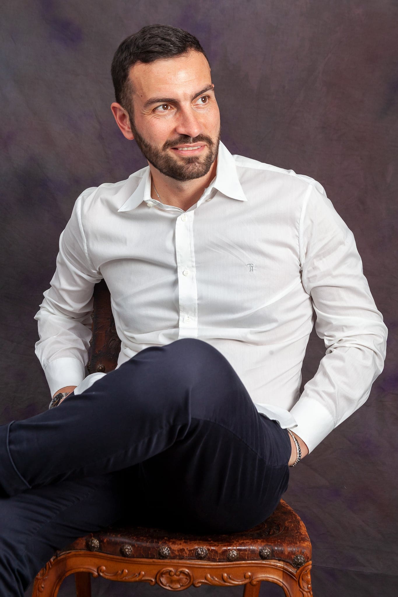 Ritratto in studio di un uomo con la barba in camicia bianca e pantaloni blu, seduto di traverso su una sedia intagliata, su fondo sfumato