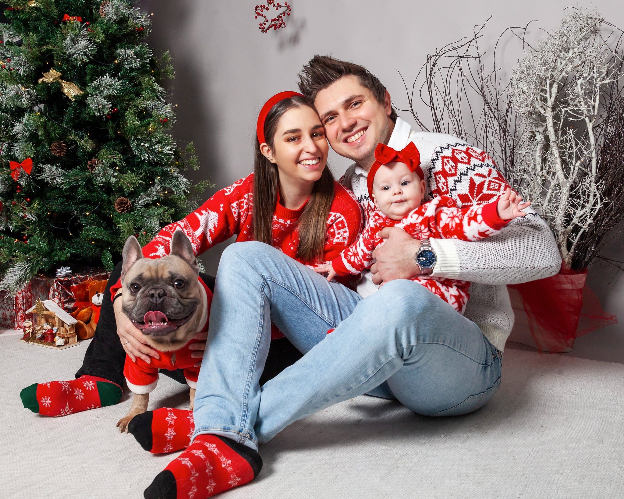 Mamma, papà, neonata e un bulldog francese in maglioni rossi e bianchi nordici, seduti a terra davanti all'albero e ai regali