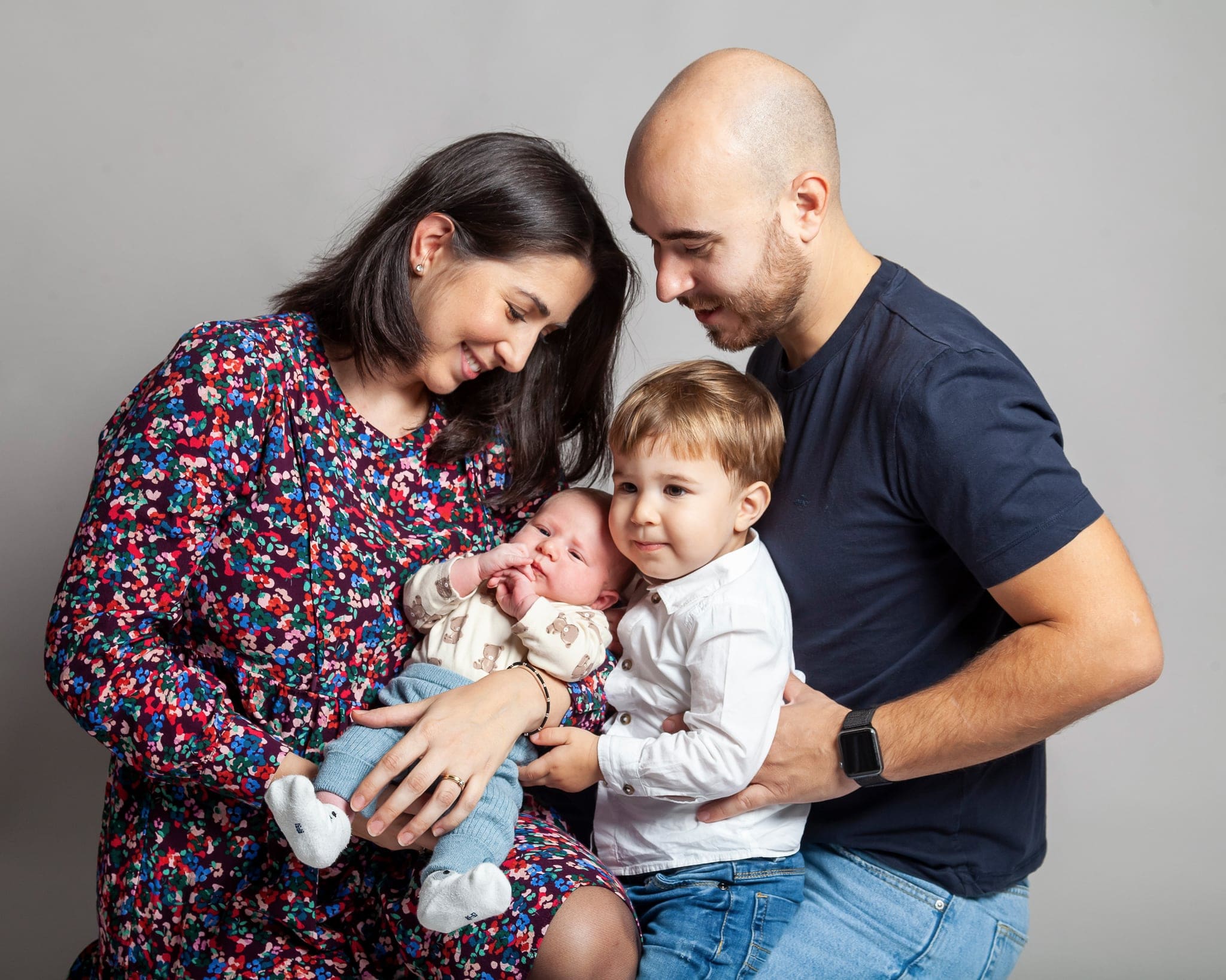 Famiglia in studio su sfondo grigio: mamma e papà con un neonato in braccio e un fratellino piccolo in camicia bianca