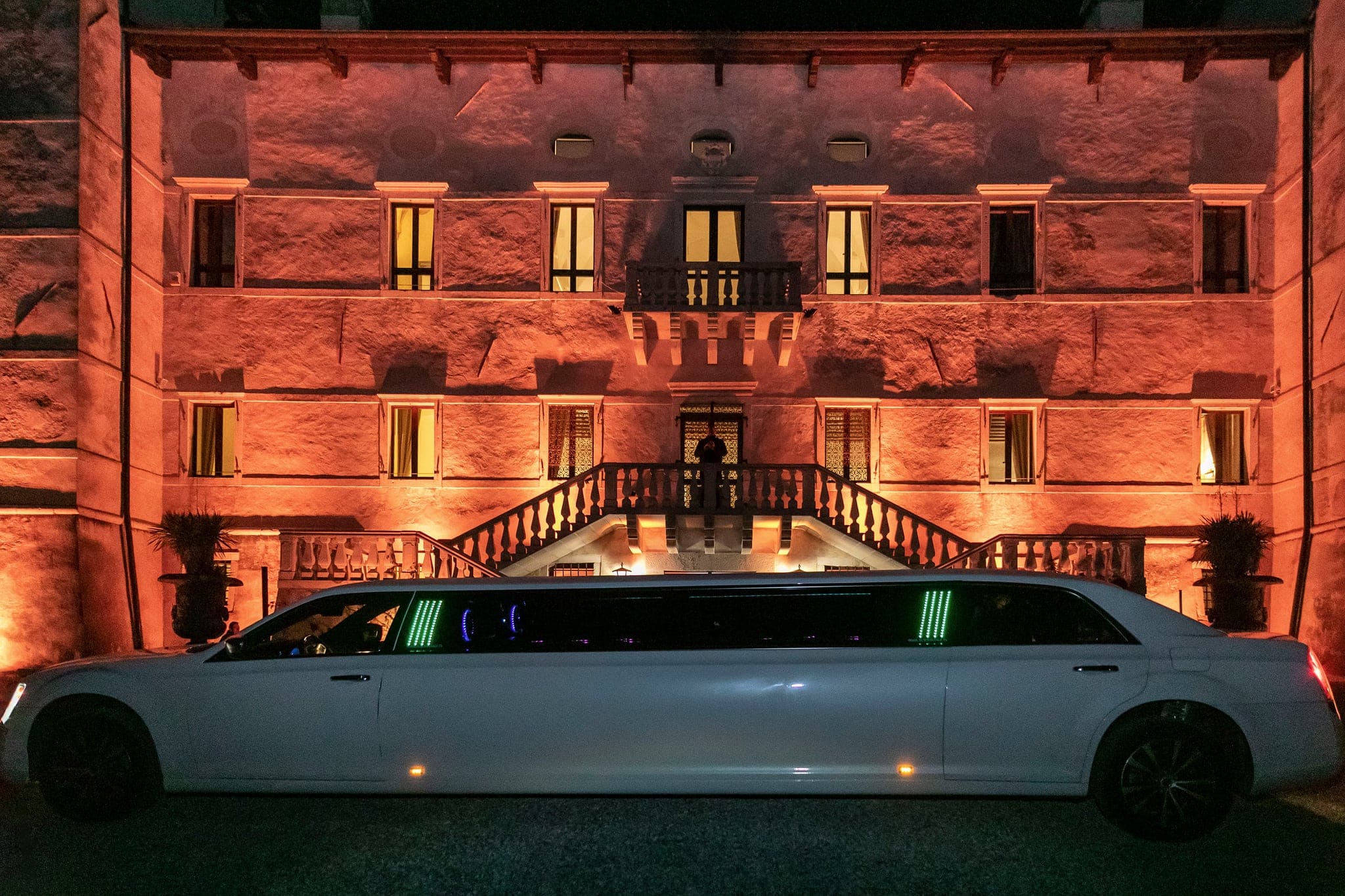 Limousine bianca parcheggiata di sera davanti a una villa storica con facciata illuminata di arancio e scalinata